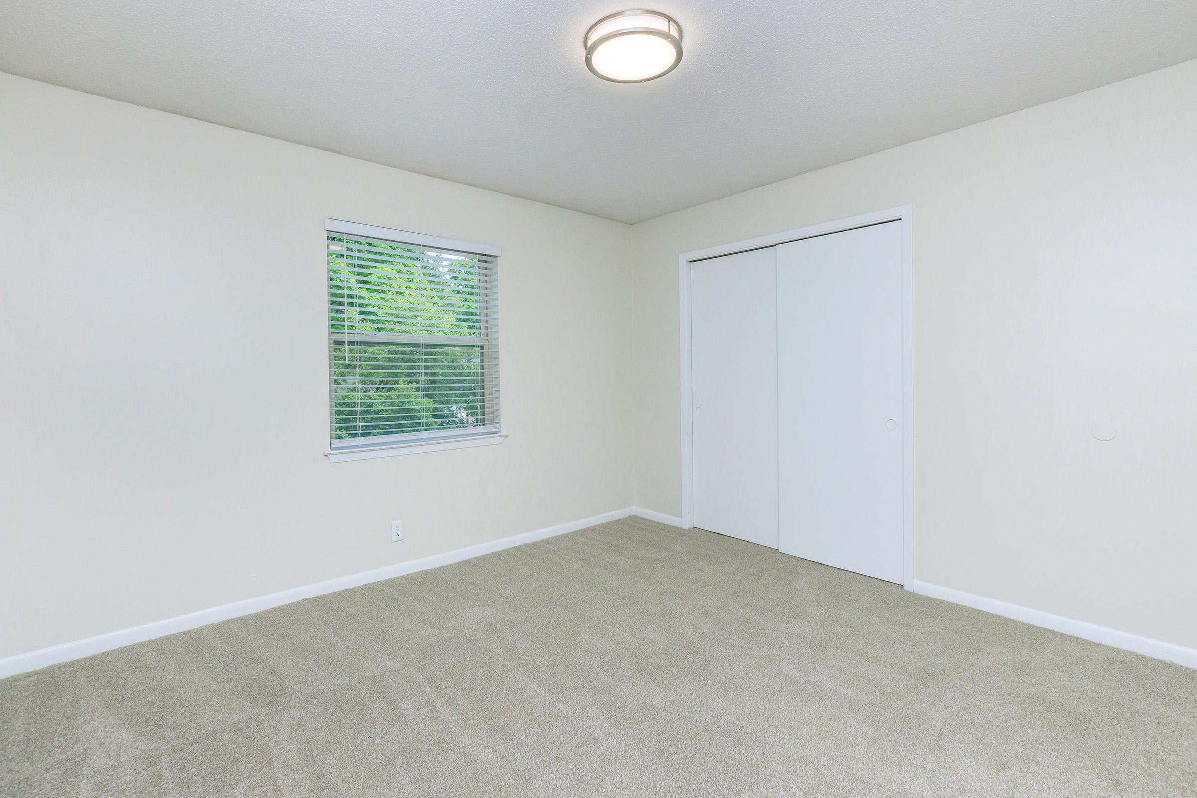 A spacious, empty room with light beige walls and a carpeted floor. A window with blinds allows natural light, and there are white double sliding doors on one wall, enhancing the room's bright, airy feel.