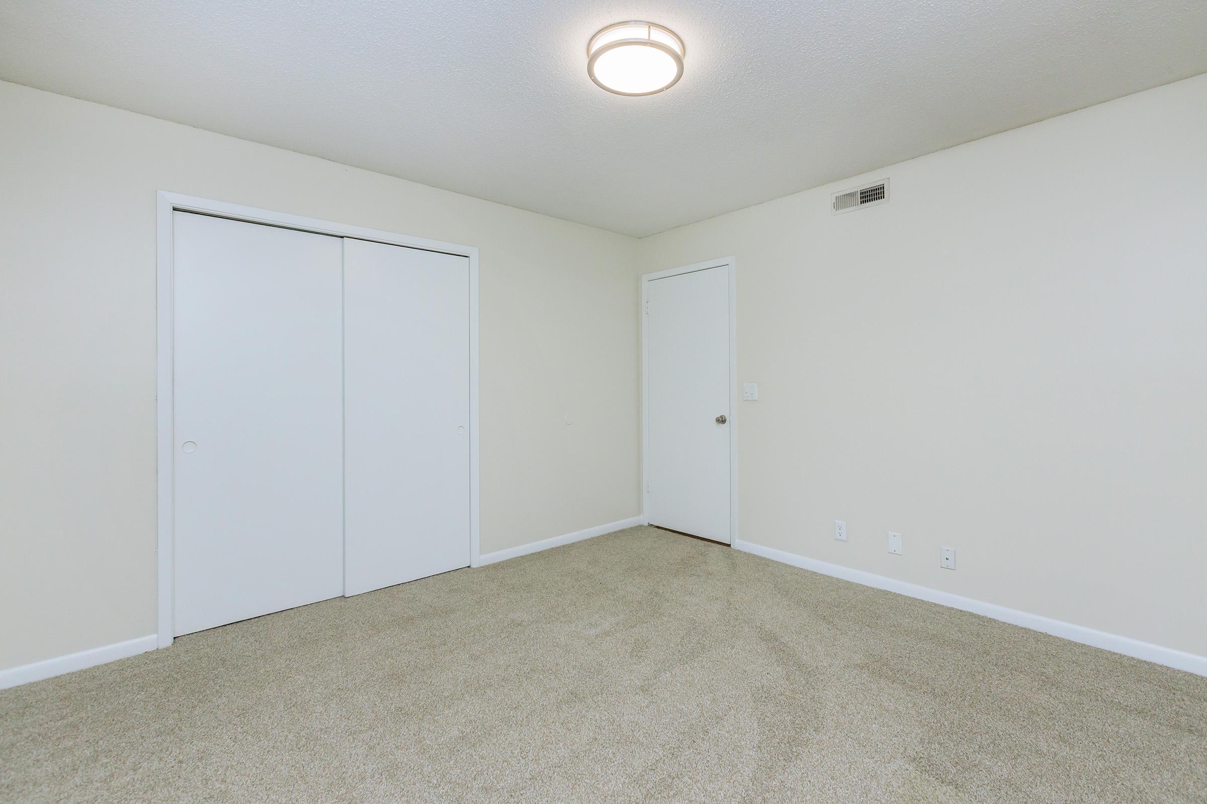 A clean, empty room featuring beige carpet, white walls, and a ceiling light. There are two sliding closet doors on one wall and a single door leading to another space. The room appears well-lit and spacious, making it suitable for various uses or decor styles.