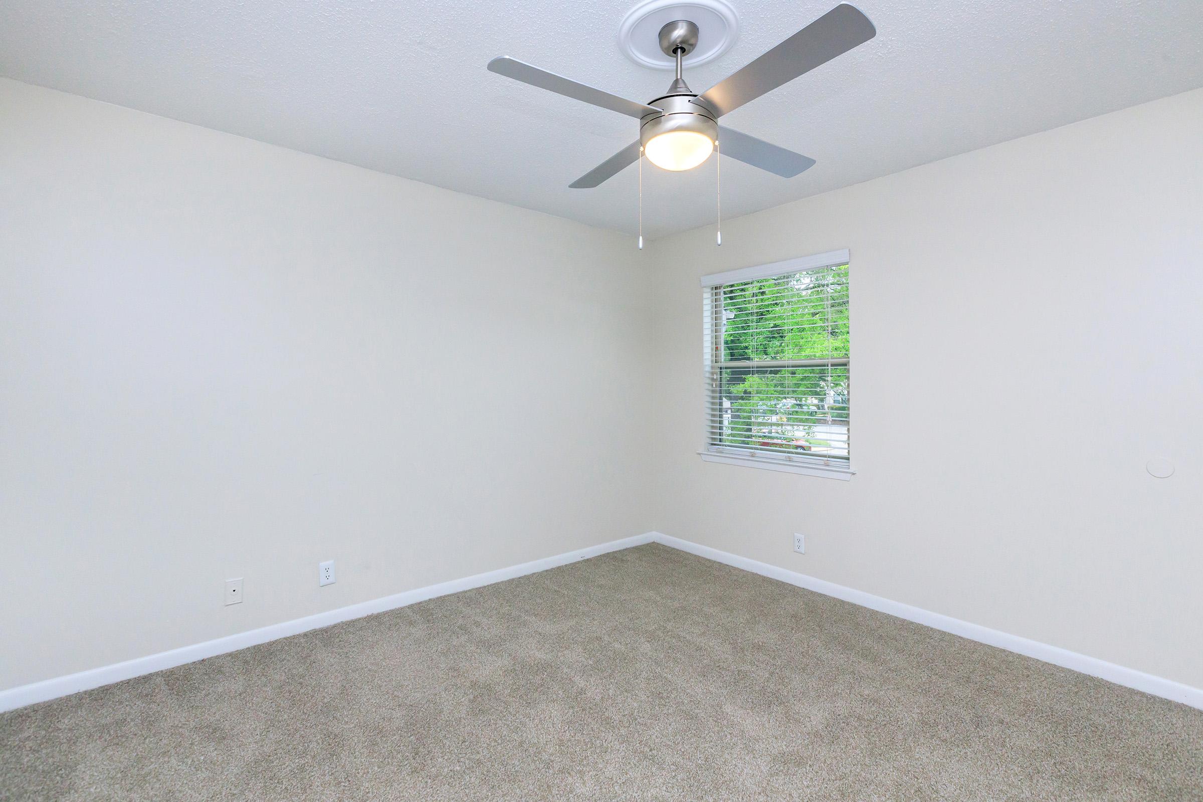An empty room with light beige walls and a ceiling fan. The floor is covered in light brown carpet. A window with blinds lets in natural light, revealing greenery outside. The space is simple and uncluttered, creating a neutral and inviting atmosphere.