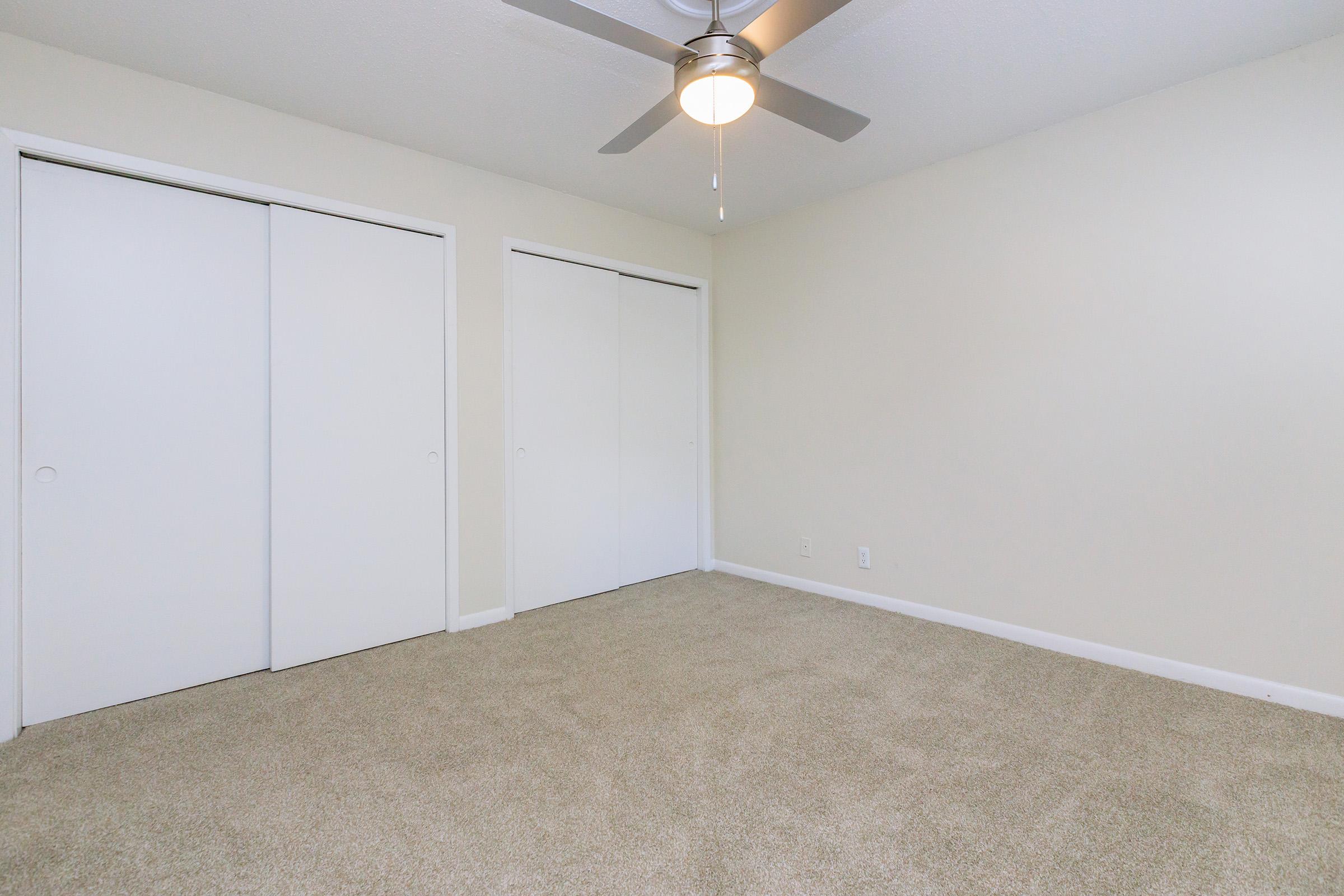 A bright and empty bedroom featuring light beige walls, light brown carpet, a ceiling fan, and two white sliding closets. The space has a clean and simple aesthetic, suitable for personal decoration.