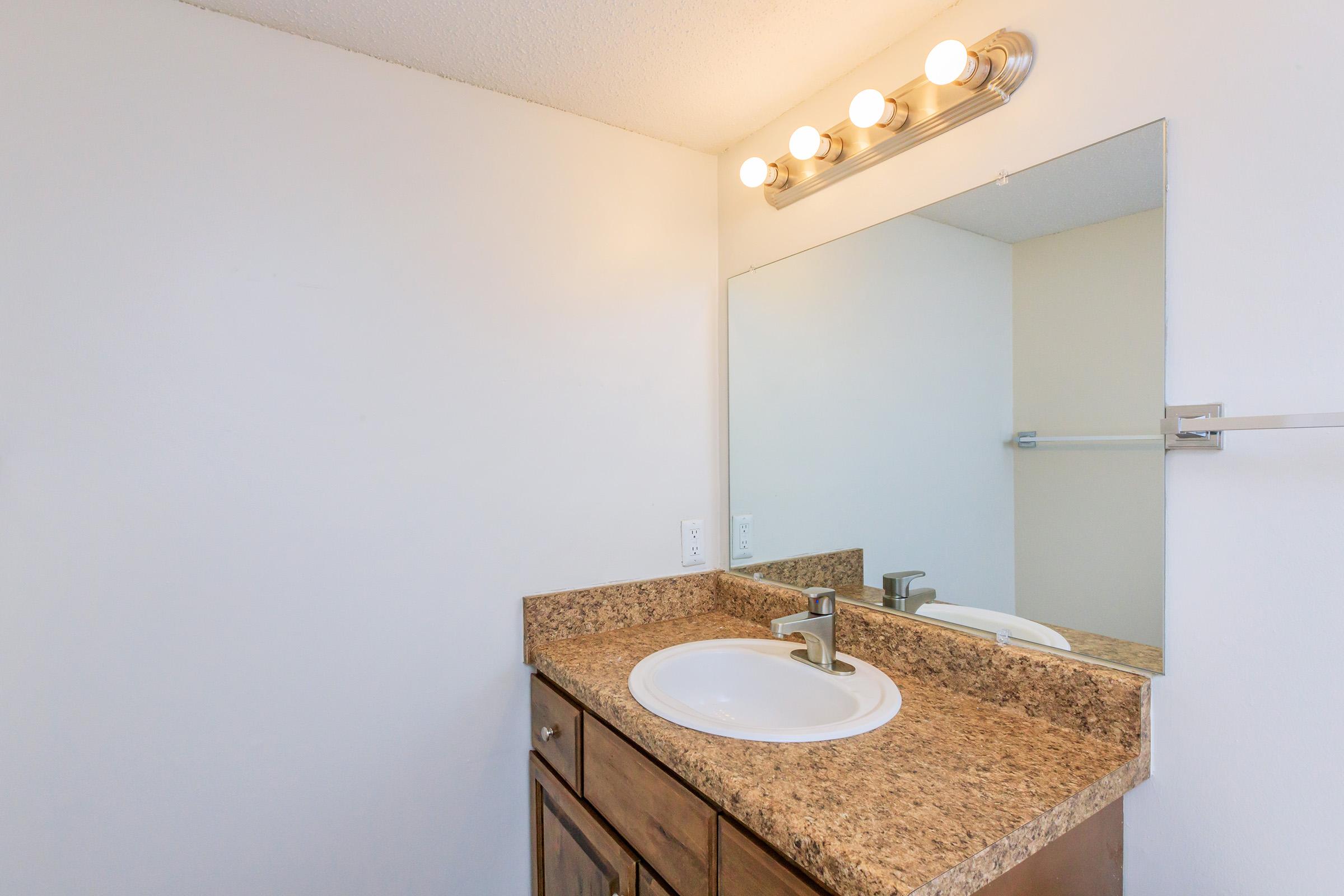 A clean bathroom vanity with a mounted mirror, light fixtures above, and a sink on a brown granite countertop. The walls are white, and there is a towel bar visible in the background. The cabinetry below the sink is made of wood, providing a simple and functional design.
