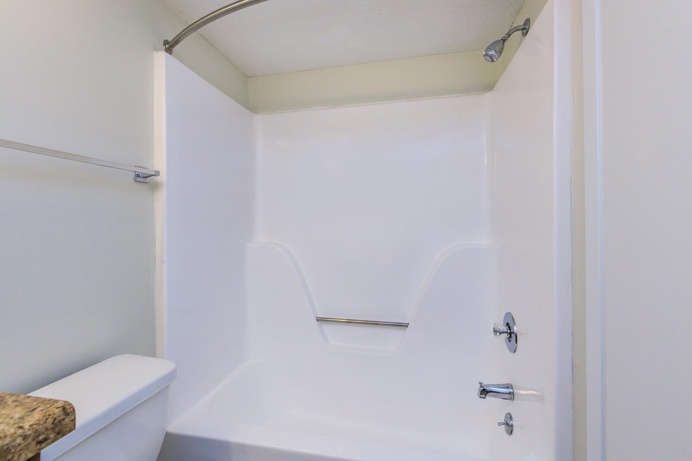 A clean, modern bathroom featuring a white acrylic bathtub and shower combination with a curved shower rod. The wall above the tub is plain white, and there is a granite countertop visible to the left, complemented by a silver faucet and showerhead.