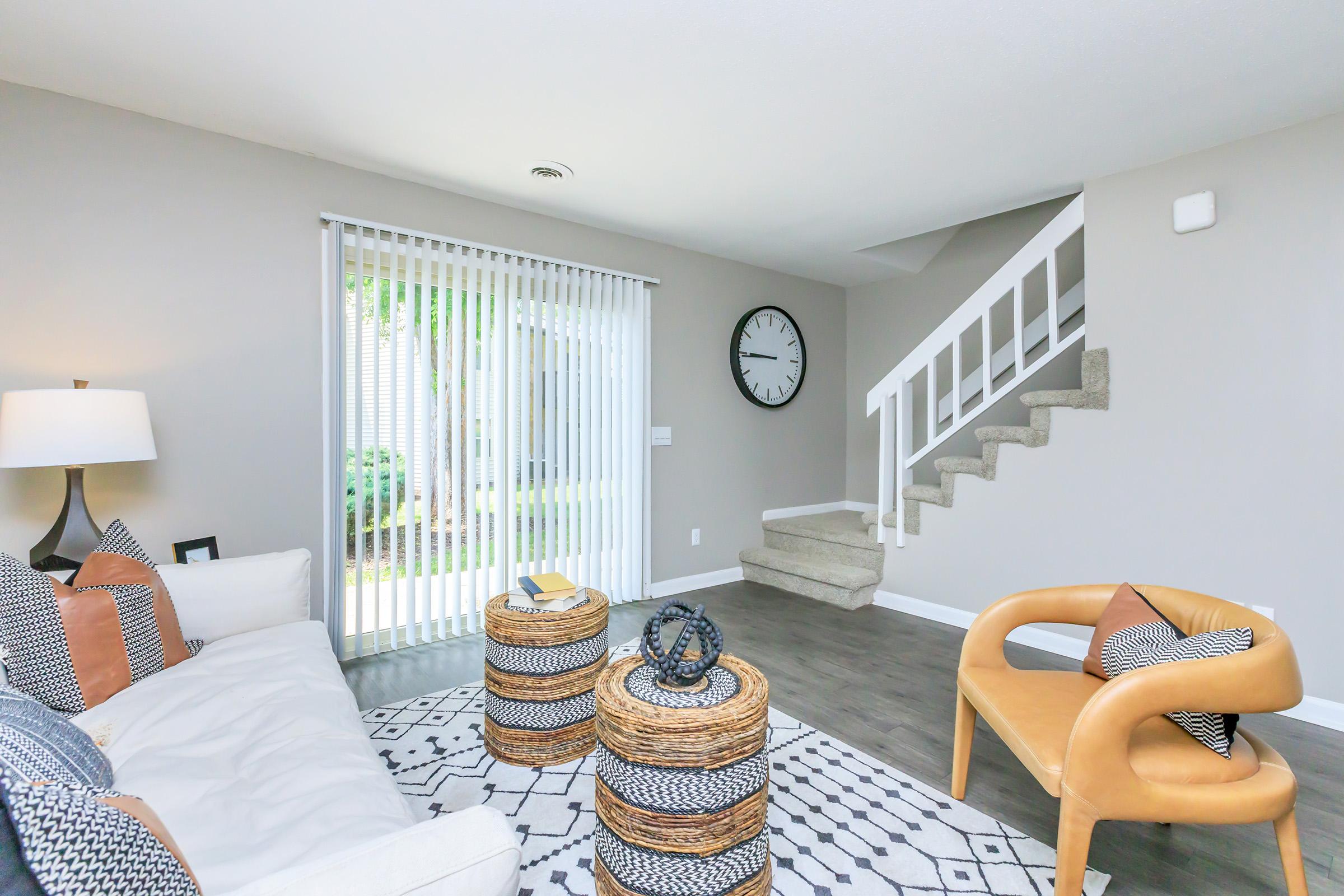 A modern, cozy living room featuring a light-colored sofa with patterned cushions, round woven coffee tables, and a stylish chair. A decorative clock hangs on the wall, and large windows with vertical blinds allow natural light to fill the space. A staircase leads to an upper level.