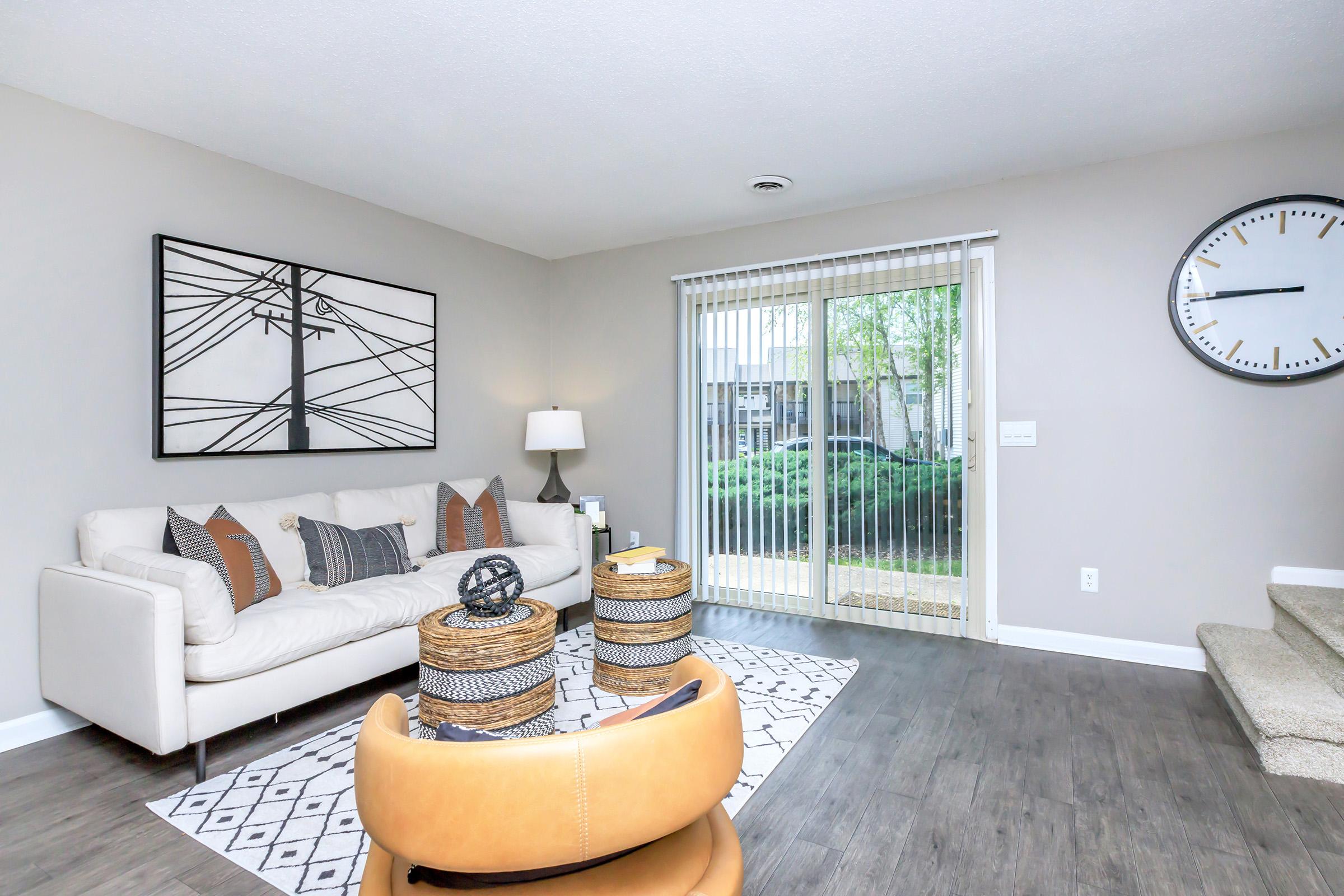 A modern living room featuring a light-colored sofa with decorative pillows, a round coffee table with woven baskets, and a large wall clock. Natural light comes through sliding glass doors leading to an outdoor view, with neutral wall colors and a patterned rug enhancing the cozy atmosphere.