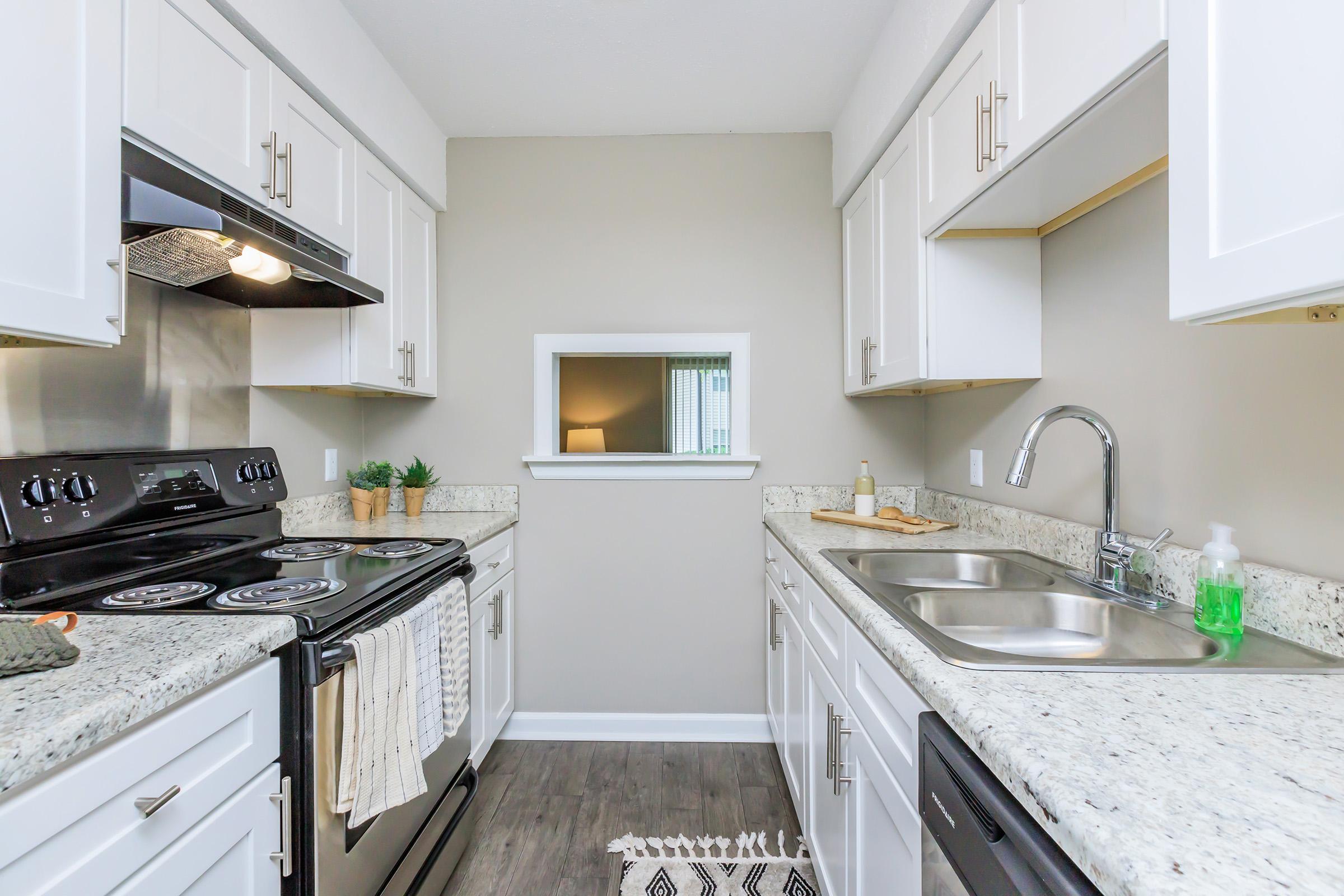 A modern kitchen featuring white cabinetry, granite countertops, a stainless steel sink, and an electric stove. The space includes a window for natural light, decorative plants on the countertop, and a patterned rug on the floor. The overall design is sleek and functional, emphasizing a clean and organized layout.