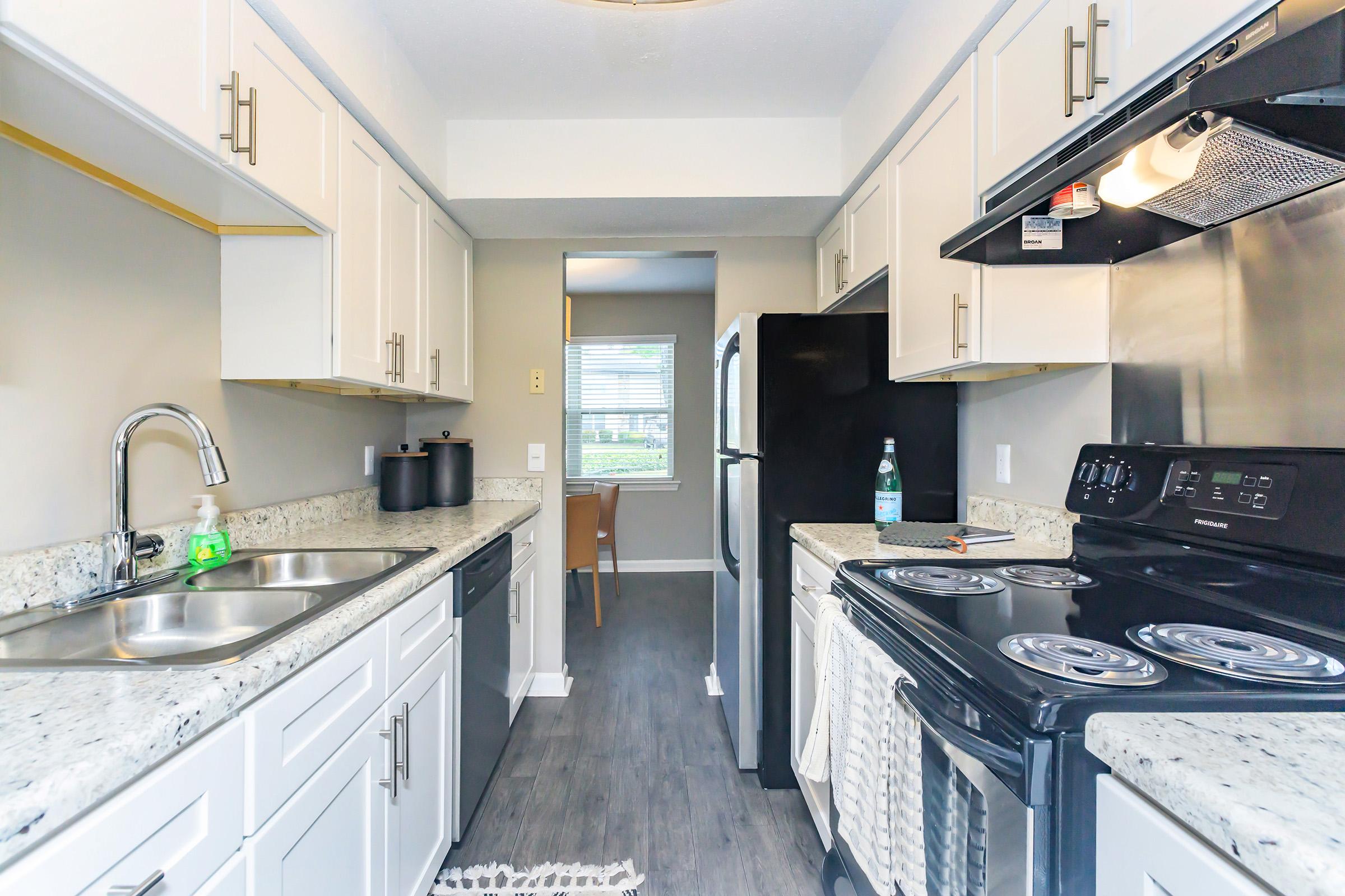 Modern kitchen featuring light-colored cabinets, granite countertops, and stainless steel appliances. Includes a double sink, an oven with stove, a dishwasher, and a black refrigerator. Natural light enters through a nearby window, creating a bright and inviting atmosphere. A small dining area is visible in the background.