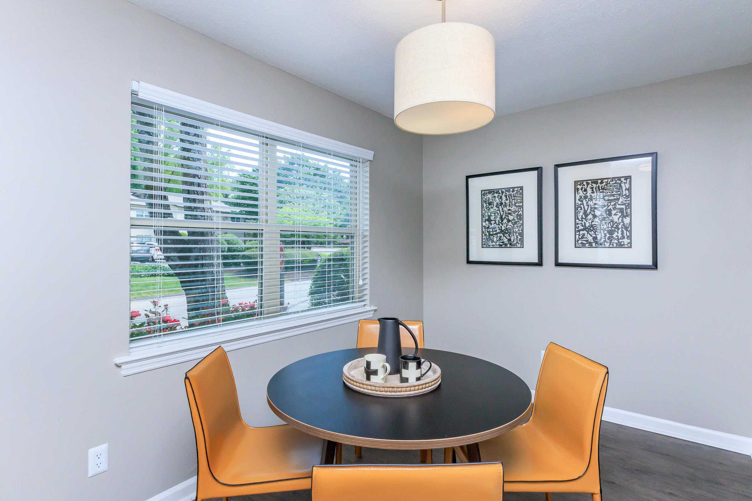 Dining area featuring a round black table surrounded by four mustard-yellow chairs. A circular pendant light hangs above the table. Two framed black-and-white artworks are displayed on the wall, and a large window with blinds allows natural light to enter, offering a view of greenery outside.