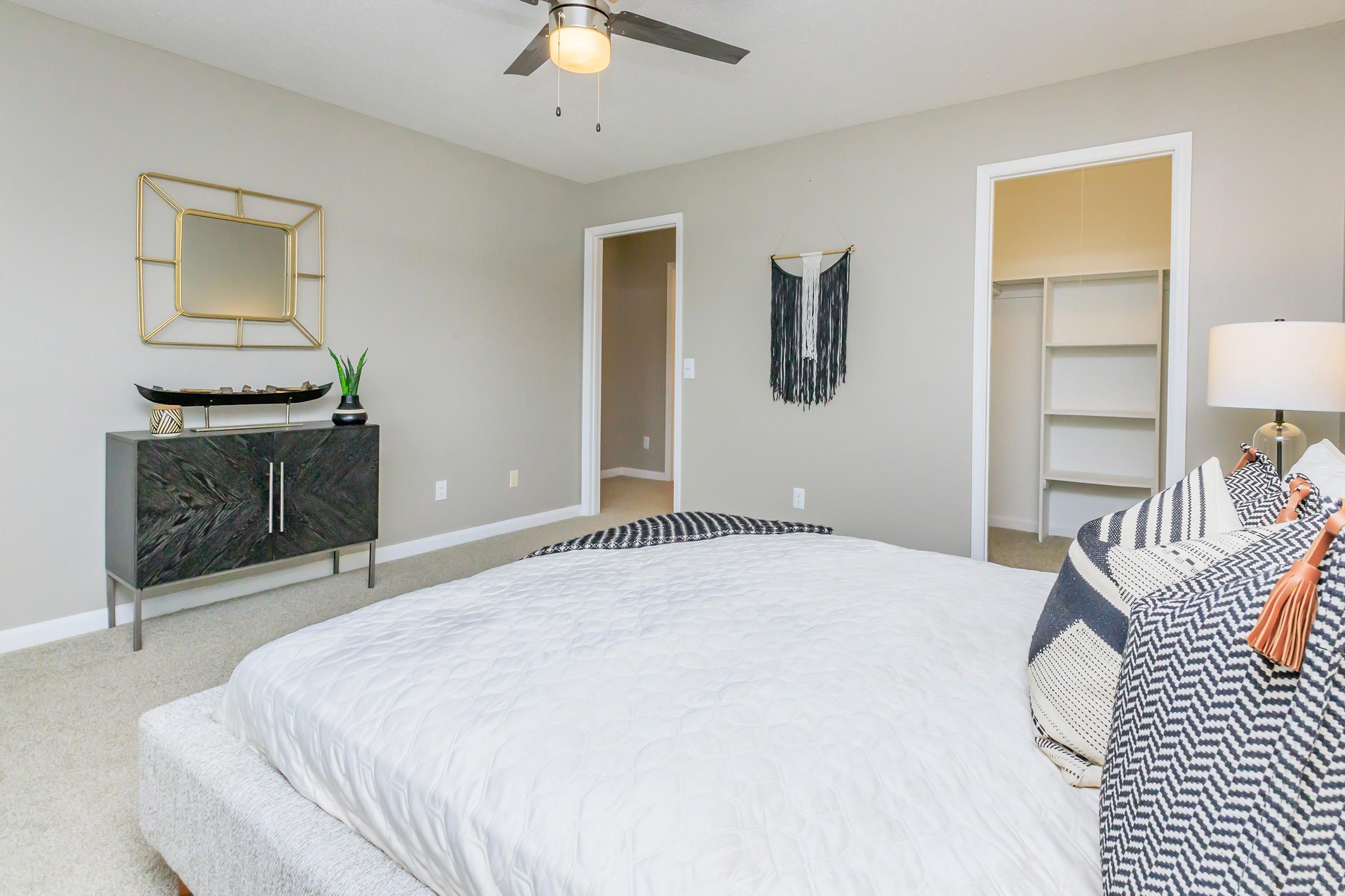 A spacious, modern bedroom featuring a neatly made bed with decorative pillows, a nightstand with a lamp, and an accent piece on the wall. A minimalist dresser and a potted plant add to the decor. The walls are painted light gray, and a walk-in closet is visible in the background, enhancing the room's inviting atmosphere.