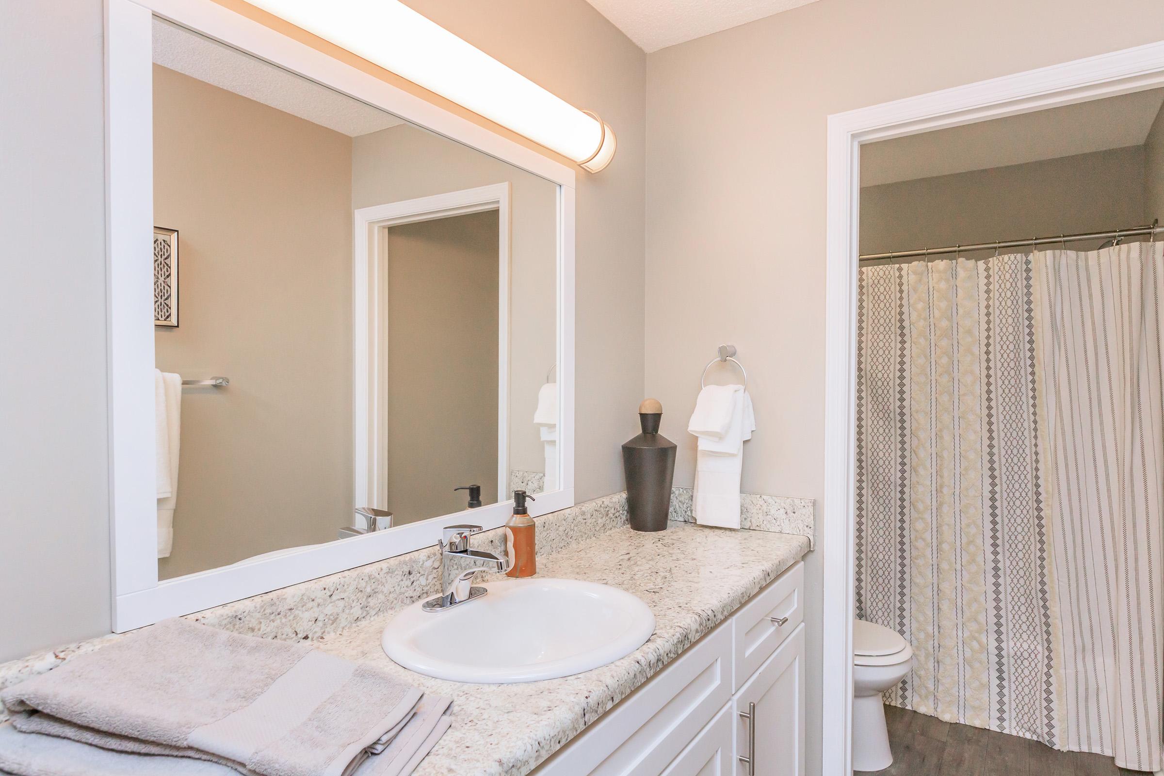 A modern bathroom featuring a granite countertop with a sink and faucet, a large mirror above, and decorative elements like a soap dispenser. White towels hang on the side, and a shower curtain with a patterned design is visible in the background. Soft lighting adds warmth to the space.