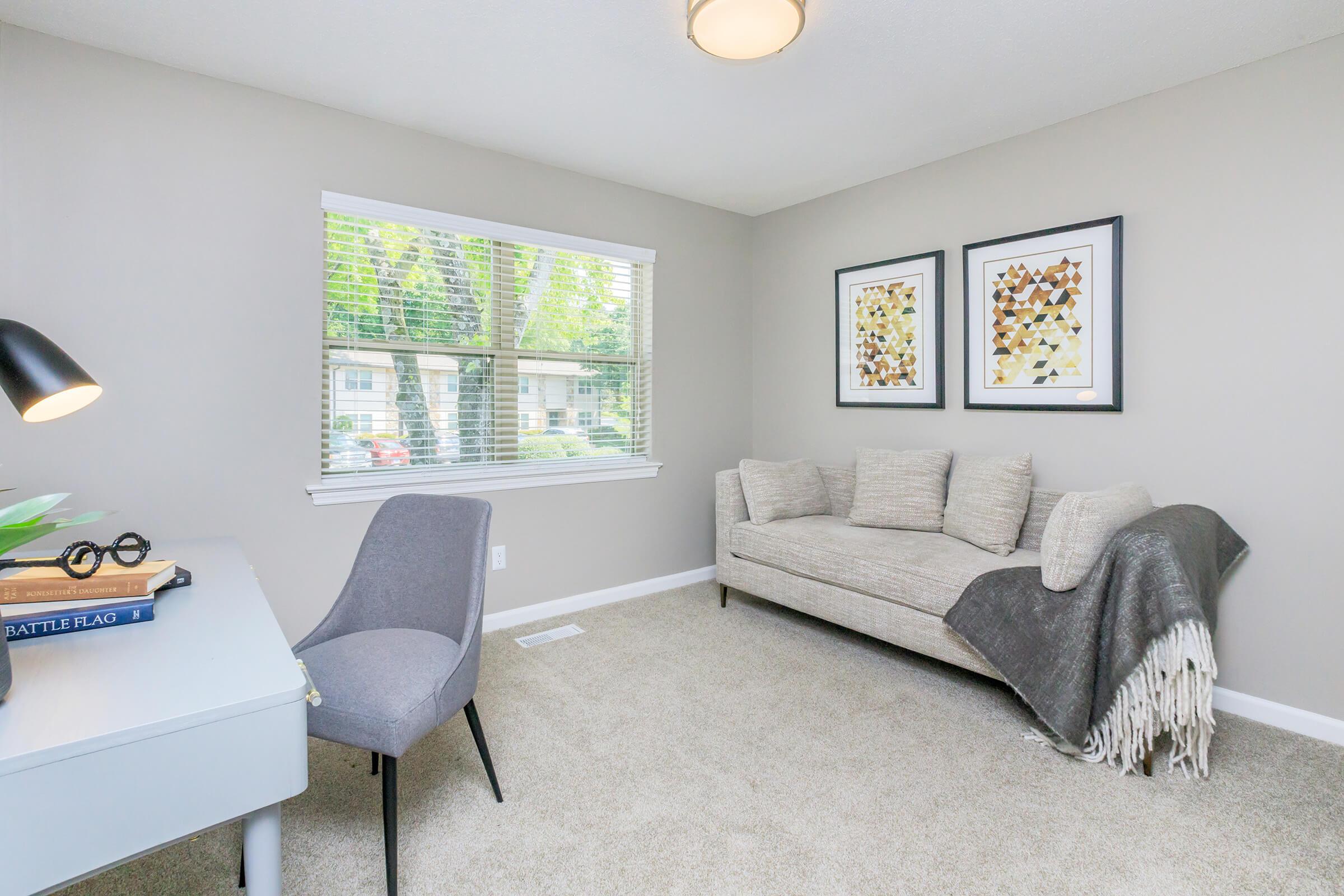 A cozy room featuring a light gray wall, a window with natural light, and a small desk with books and a lamp. There's a stylish gray couch adorned with a blanket, and two framed wall art pieces. A simple gray chair complements the modern décor, creating an inviting and comfortable atmosphere.