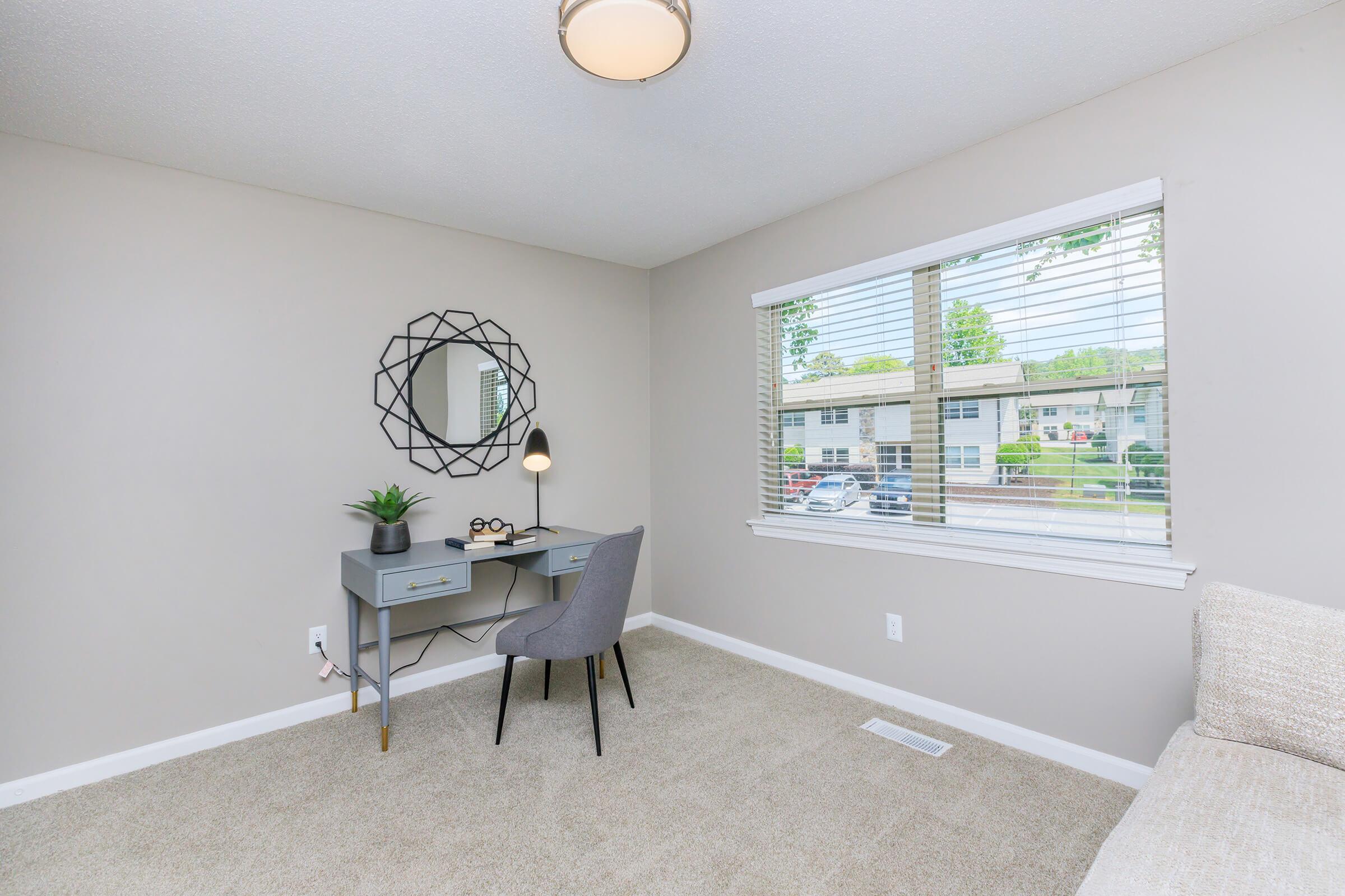 A modern, minimalist room featuring a light gray carpet and beige walls. There's a small desk with a lamp and a plant, positioned next to a window with blinds. A round mirror hangs on the wall, and a cozy sofa is partially visible to the right. Natural light brightens the space, creating a warm atmosphere.