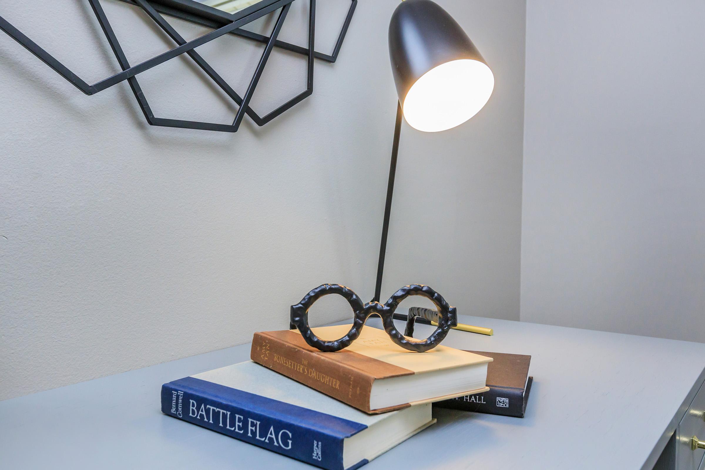 A stylish lamp illuminates a stack of books on a gray surface. The top book has a brown cover, and underneath are two blue-covered books. A pair of black, round-framed glasses rests on top, surrounded by a modern geometric mirror on the wall in the background.