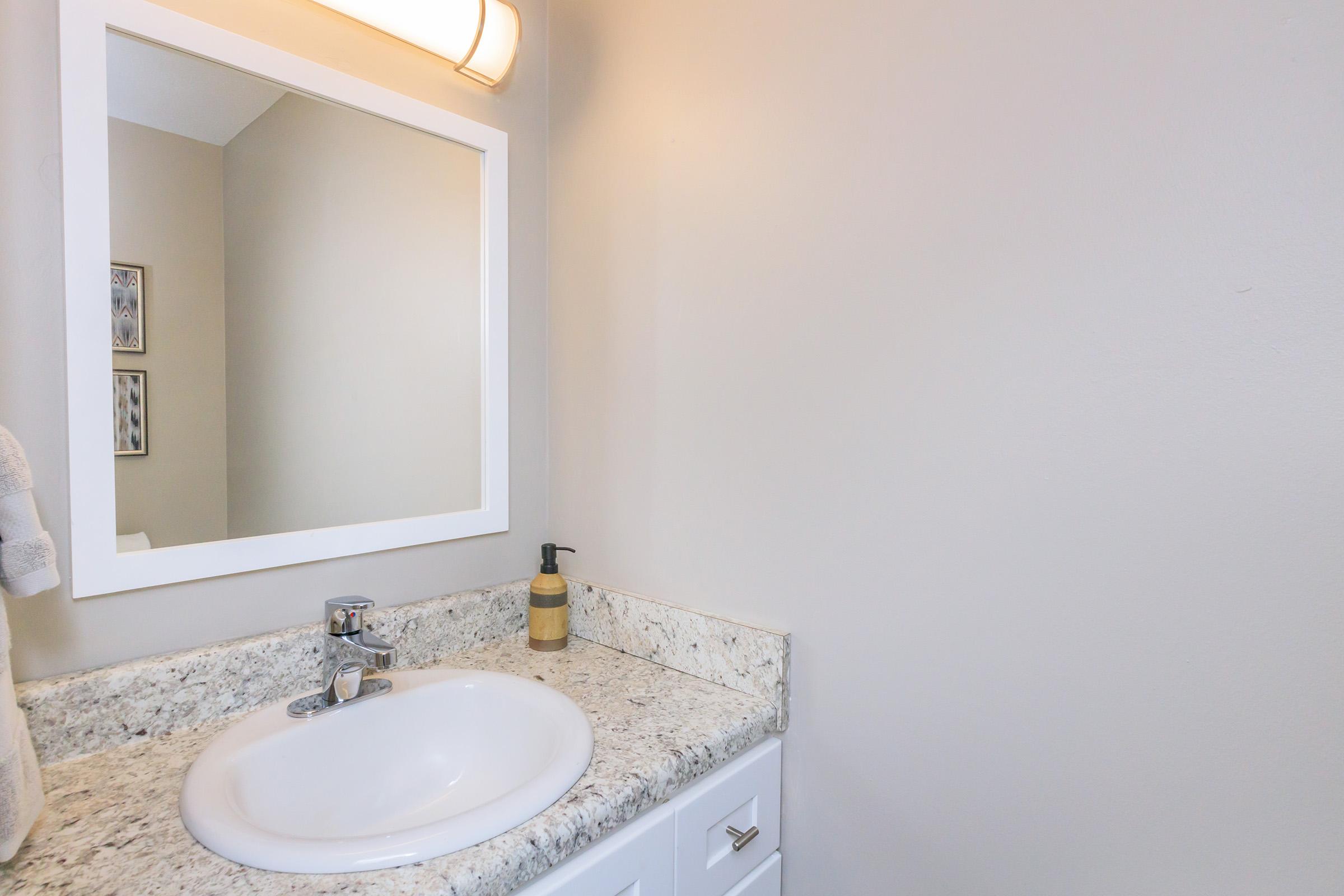 A clean bathroom sink area featuring a white countertop with a round sink, a modern faucet, and a beige soap dispenser. The wall is painted light gray, and there is a large rectangular mirror above the sink. A towel is neatly hung nearby, contributing to a tidy and minimalistic aesthetic.