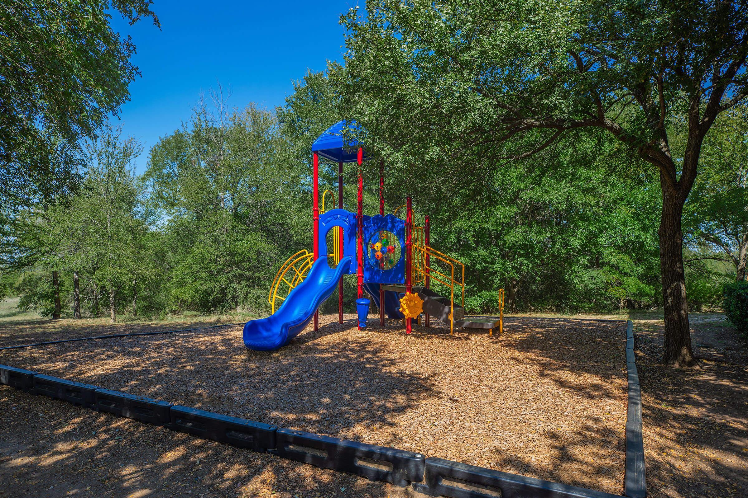 A colorful playground structure featuring a blue slide, climbing features, and a small deck, surrounded by a natural setting with trees. The ground is covered with wood chips, and the sky is clear and blue, suggesting a sunny day.