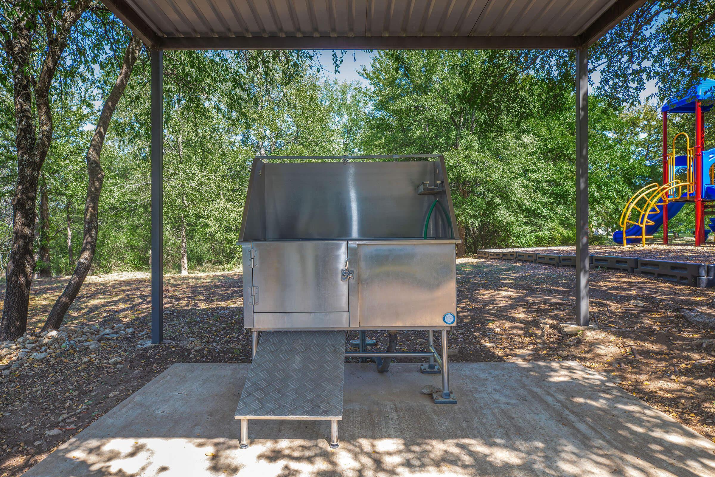 A metal waste disposal unit with an inclined ramp, located under a shelter surrounded by trees. In the background, a colorful playground with slides and climbing structures can be seen. The setting appears to be in a park or recreational area.