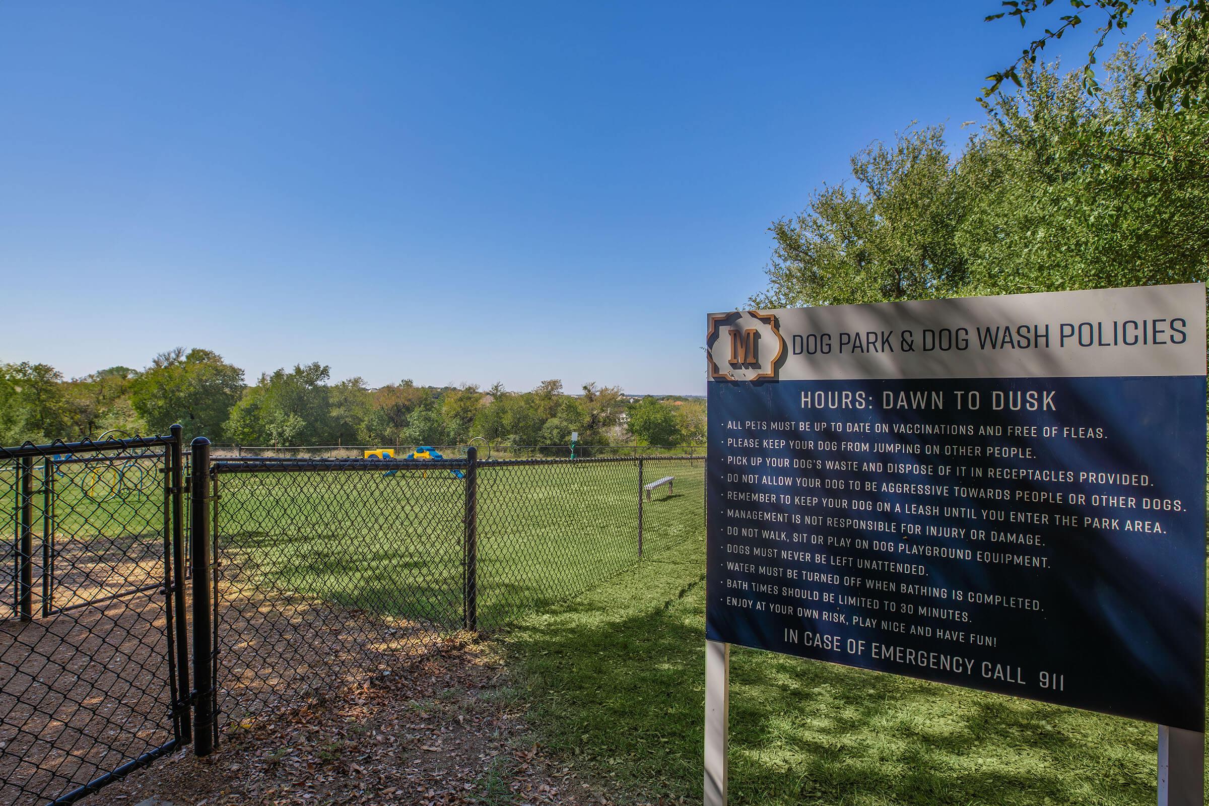 A fenced dog park with a sign detailing park policies and hours. The sign states the park is open from dawn to dusk and lists rules for dog owners, including vaccination requirements. In the background, there are trees and a play area for dogs.