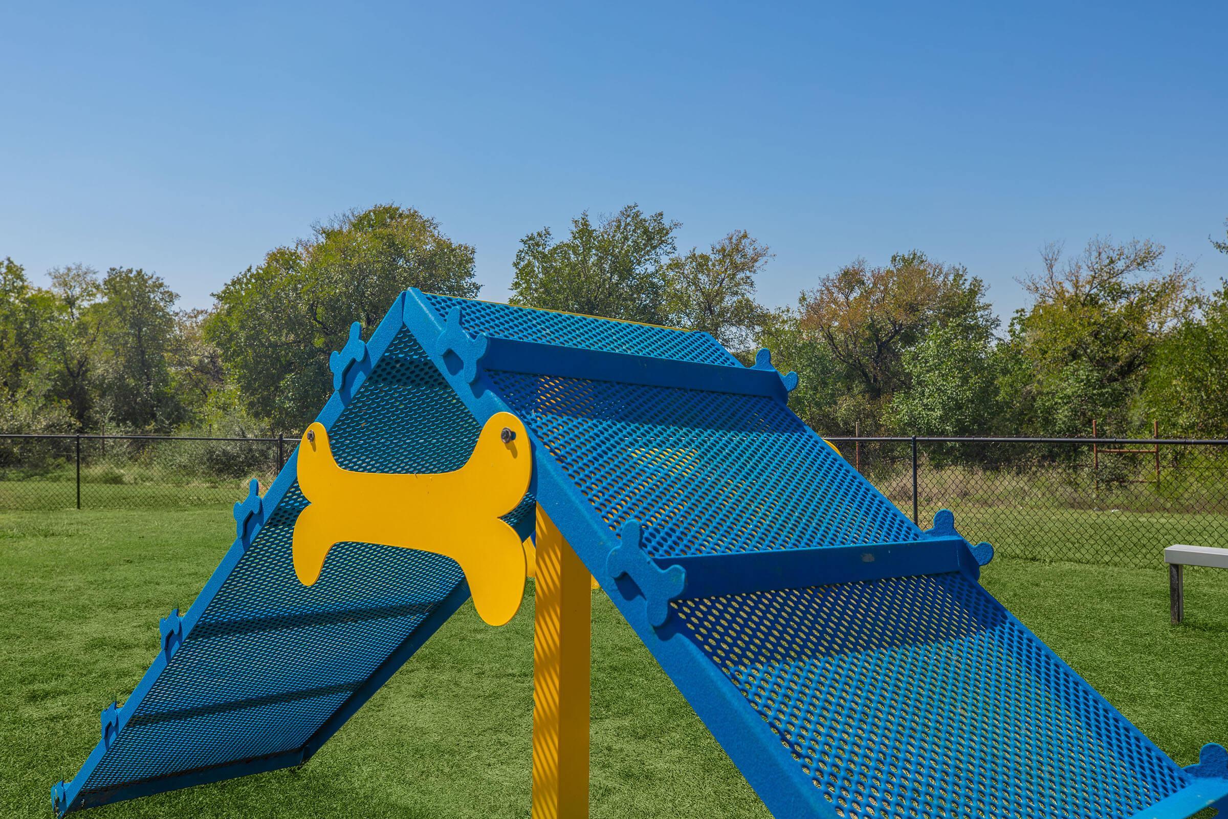 A blue dog agility ramp with a yellow dog silhouette design, set in a grassy area with a clear blue sky and scattered trees in the background. The ramp features a textured surface for traction and is designed for training dogs in an outdoor play space.
