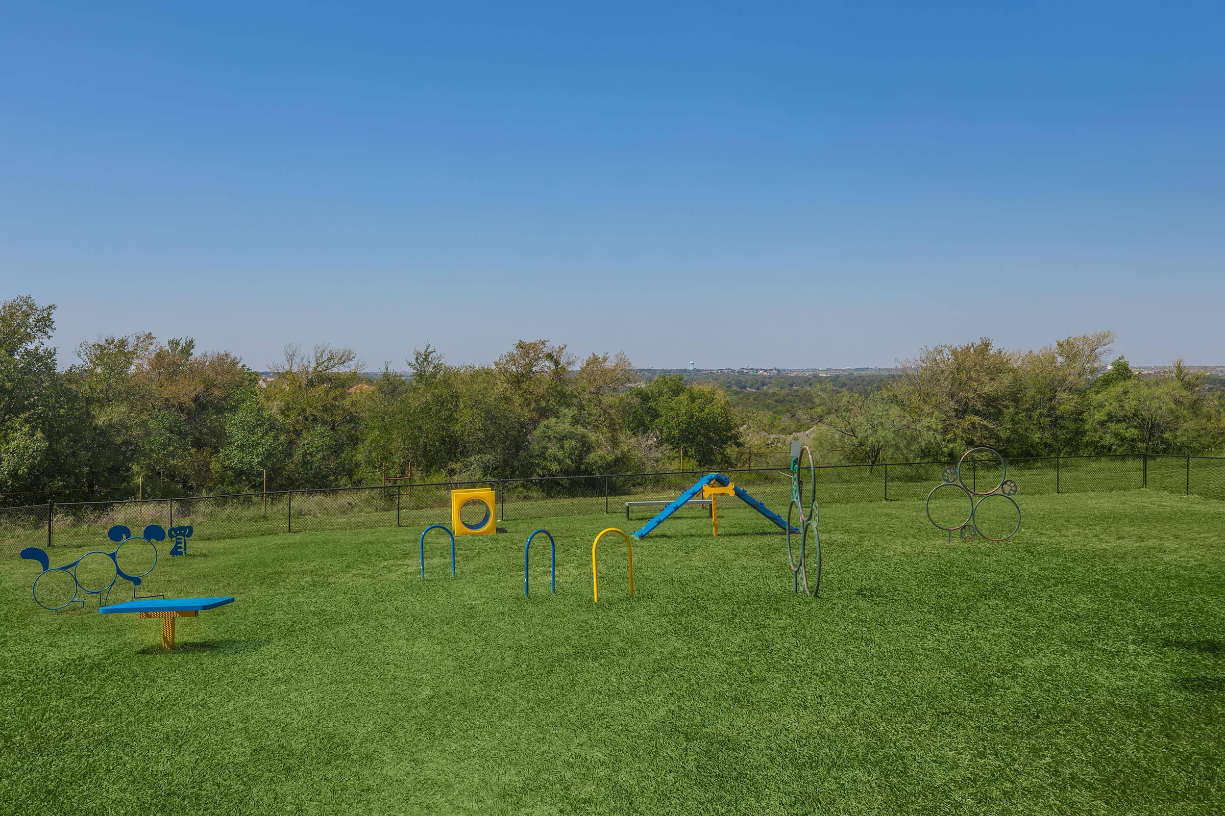 A colorful outdoor playground with various play structures, including a slide, climbing frame, and balance elements, set on a grassy area. The background features trees and a clear blue sky, creating a bright and inviting atmosphere for children to play and explore.