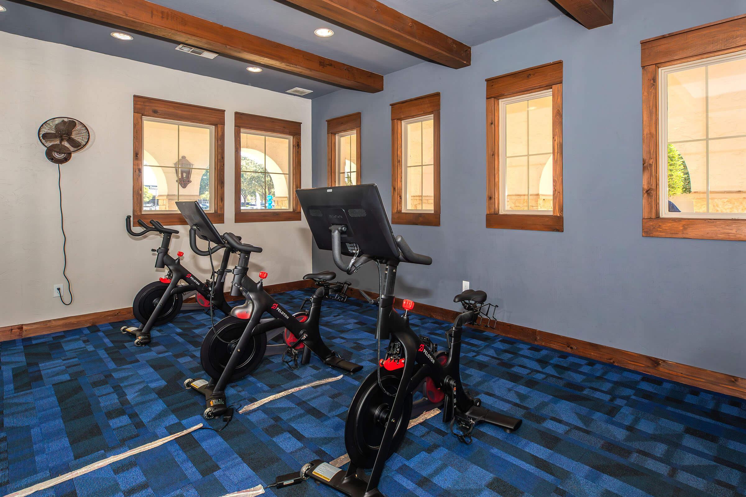 A bright and spacious indoor cycling room featuring three stationary bikes on a blue carpet. Large windows provide natural light, and a wall-mounted fan is visible in the corner, enhancing the workout environment. The room has wooden beams and a modern design, creating an inviting space for exercise.