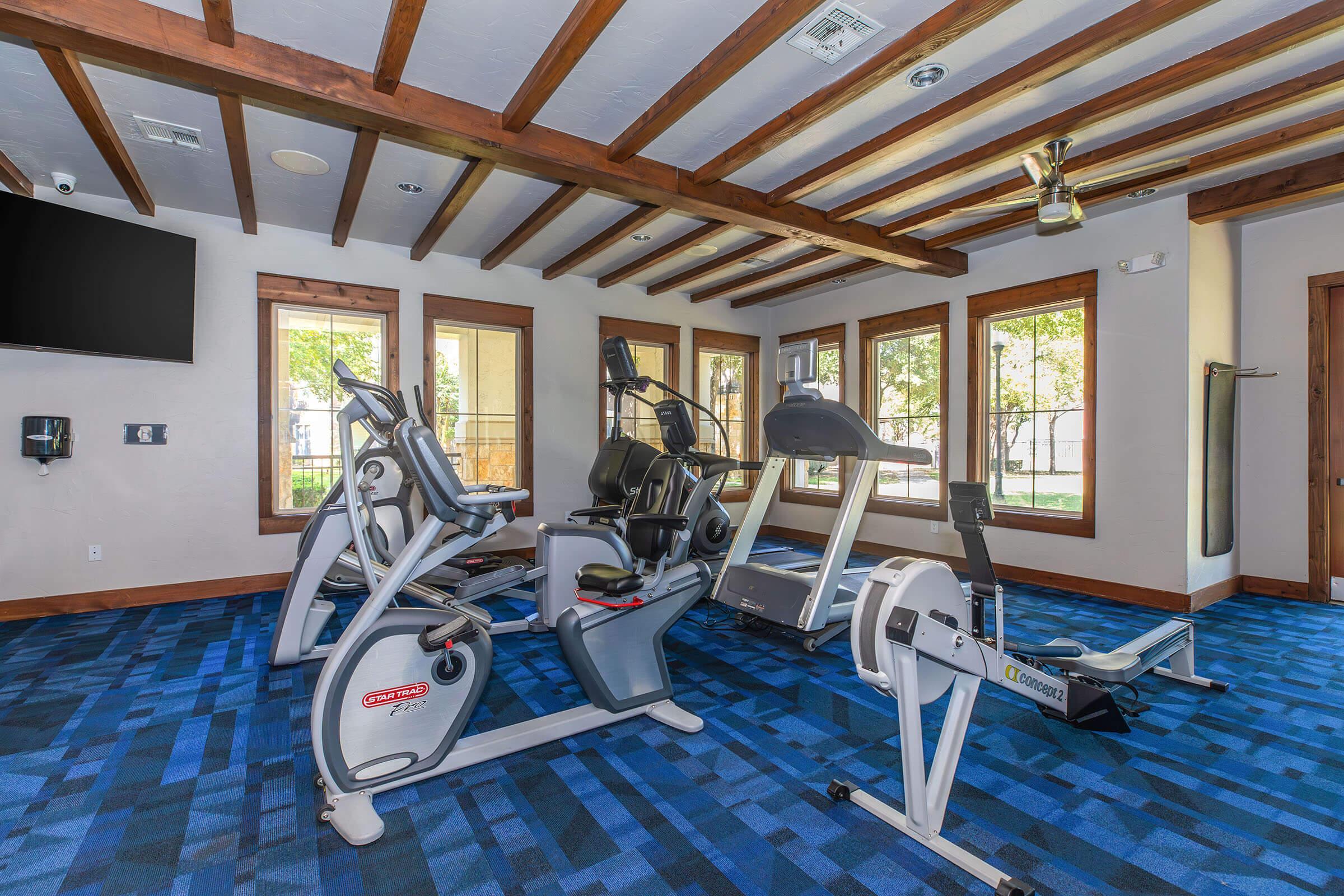 A modern fitness room featuring exercise equipment including a stationary bike, elliptical machine, and rowing machine. The space has a blue carpet, wooden beam ceiling, and large windows allowing natural light to fill the room. A flat-screen TV is mounted on the wall.