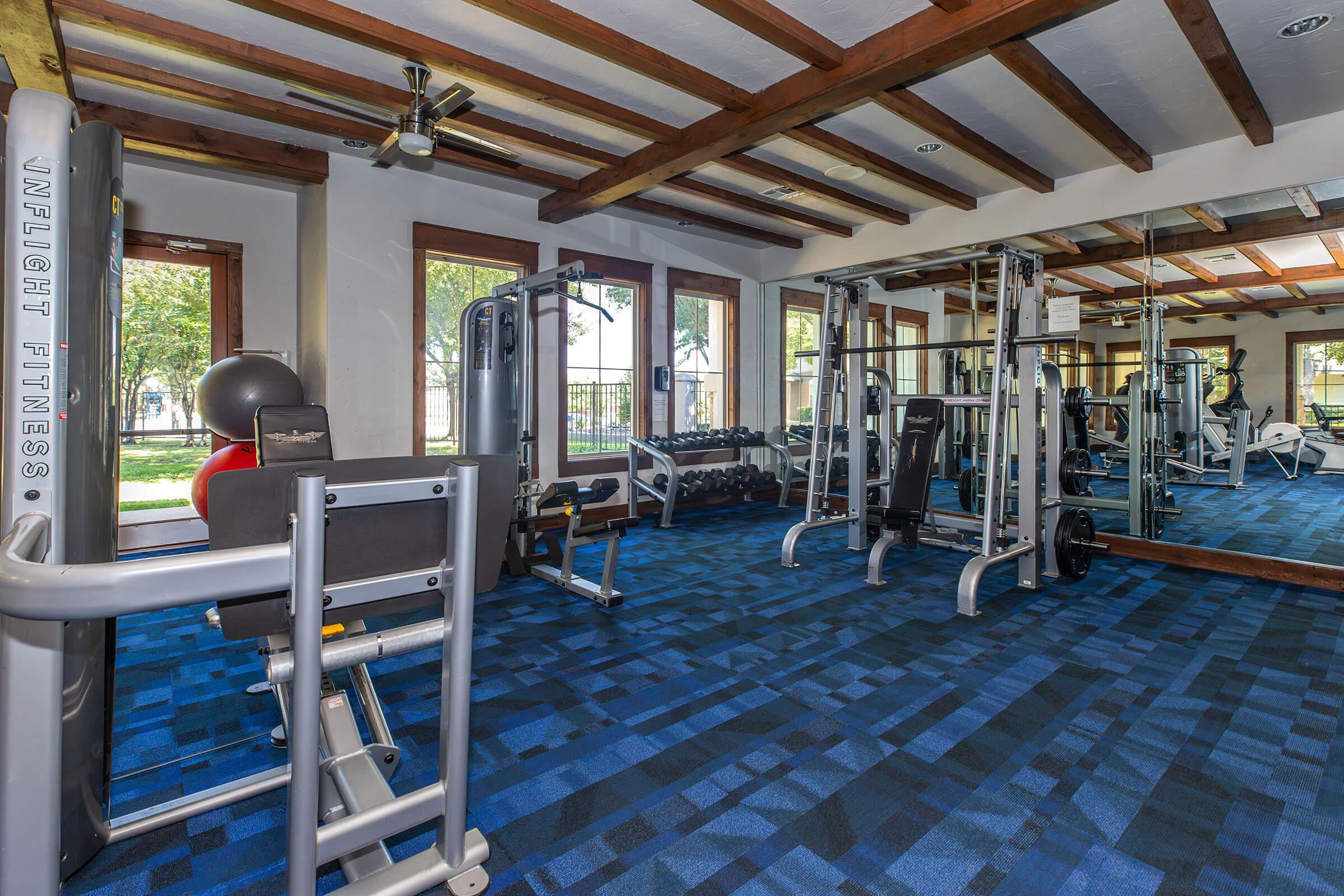 A modern gym interior featuring various fitness equipment, including strength training machines, free weights, and exercise mats. The space has large windows providing natural light, a blue carpet, and wooden beams on the ceiling, creating an inviting atmosphere for workouts.