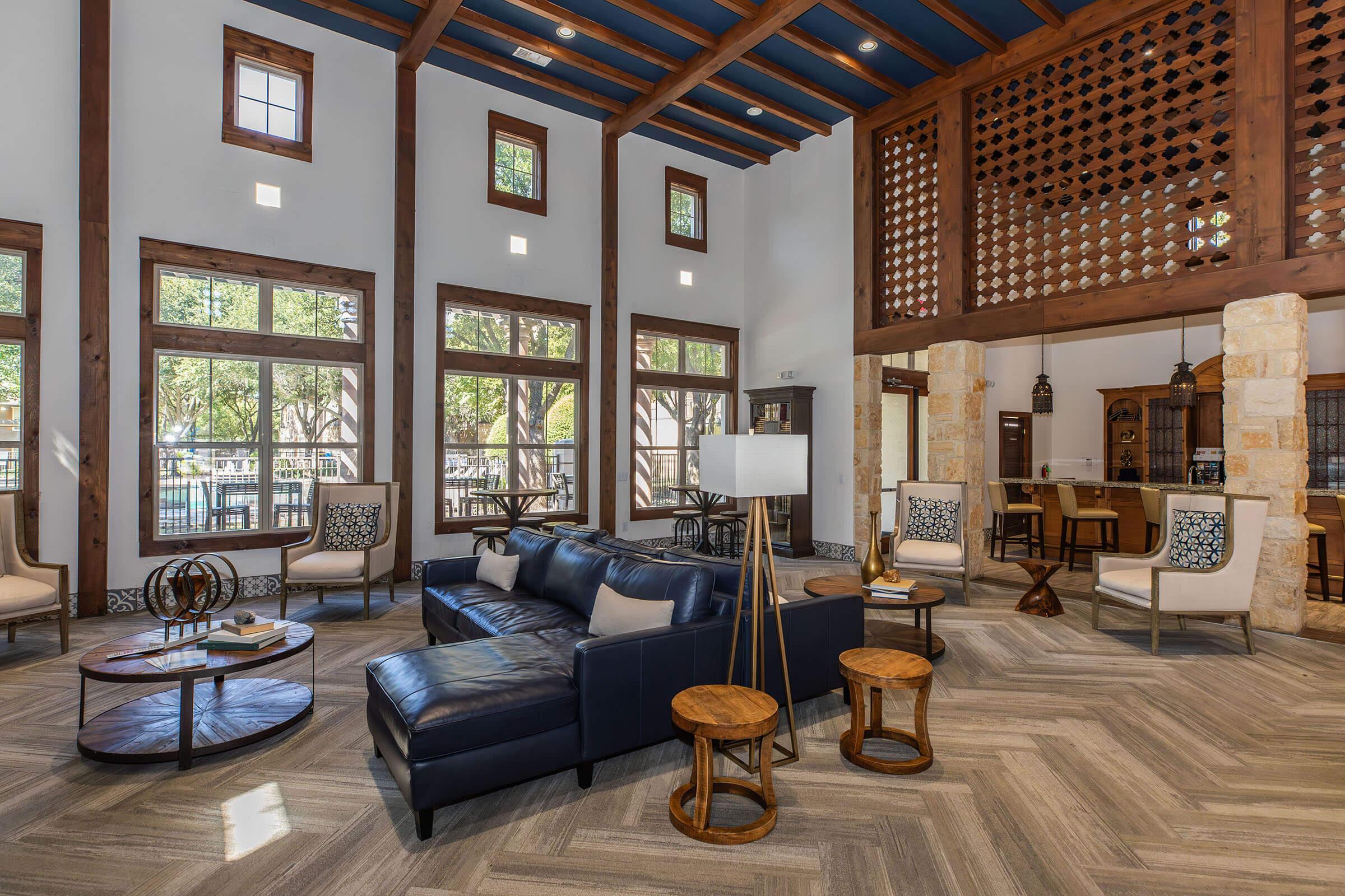 Interior of a modern living space featuring a dark leather sectional sofa, wooden coffee tables, and stylish chairs. Large windows allow natural light to fill the room, highlighting the wooden beams and decorative accents. A kitchenette is visible in the background, enhancing the inviting atmosphere.