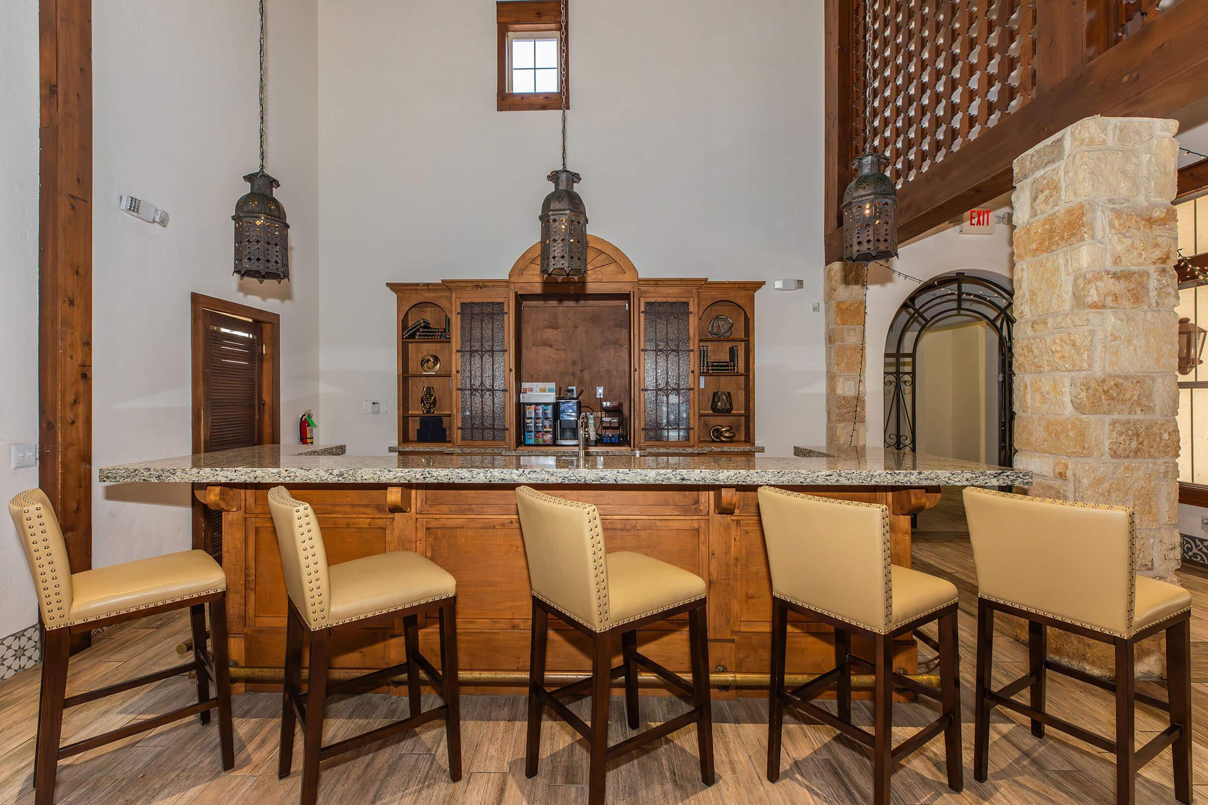 A rustic bar area featuring a wooden counter and beige bar stools. The backdrop includes a decorative wooden cabinet with various items displayed. Pendant lights hang from the ceiling, illuminating the space, which features light-colored stone walls and wooden beams. Natural light filters in through a window.