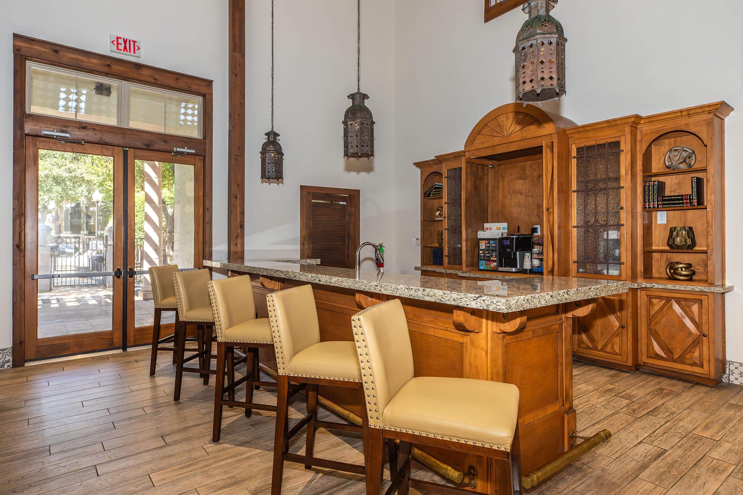 Interior of a modern lobby featuring a wooden reception desk with a granite countertop, several beige upholstered bar stools, and decorative lanterns hanging from the ceiling. Large windows allow natural light and there are shelves with books and decorative items.