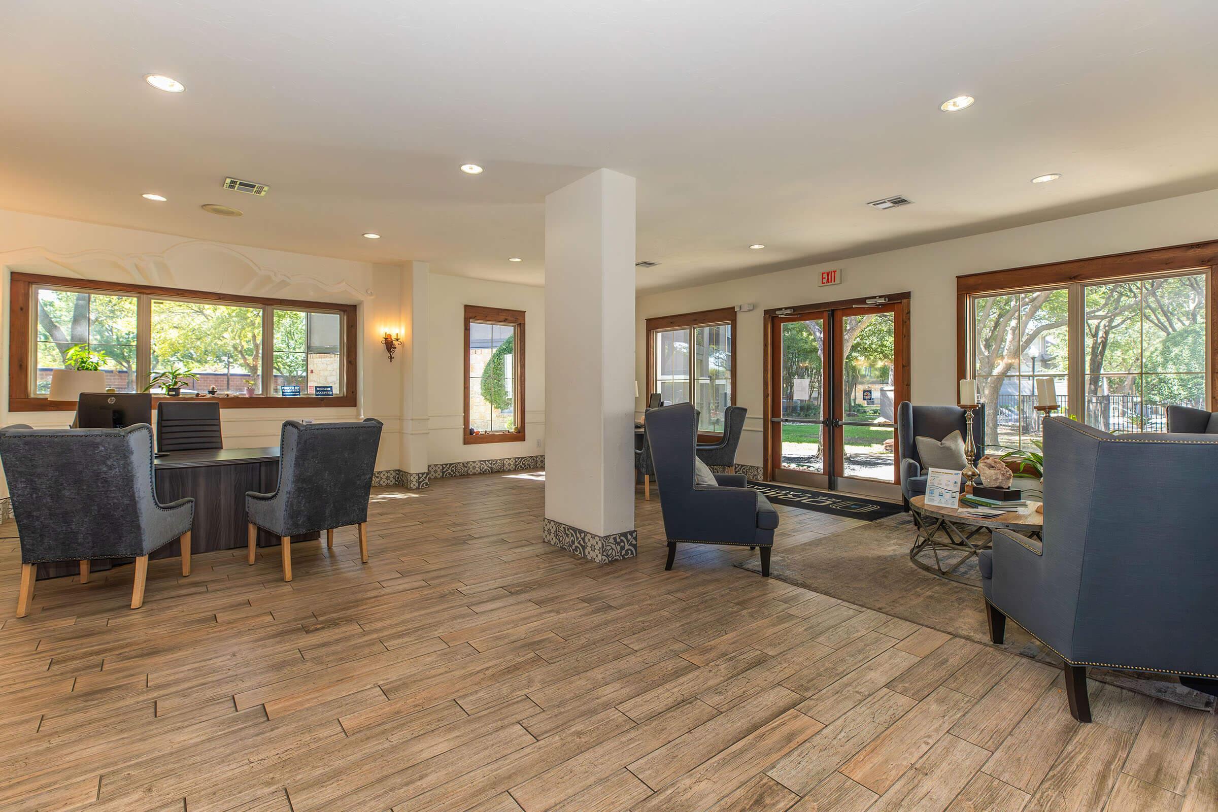 A spacious lobby area with large windows allowing natural light. Comfortable seating arrangements with dark blue chairs and a dining table. Wooden flooring and decorative lighting create a welcoming atmosphere. The entrance features double doors that open to an outdoor view.