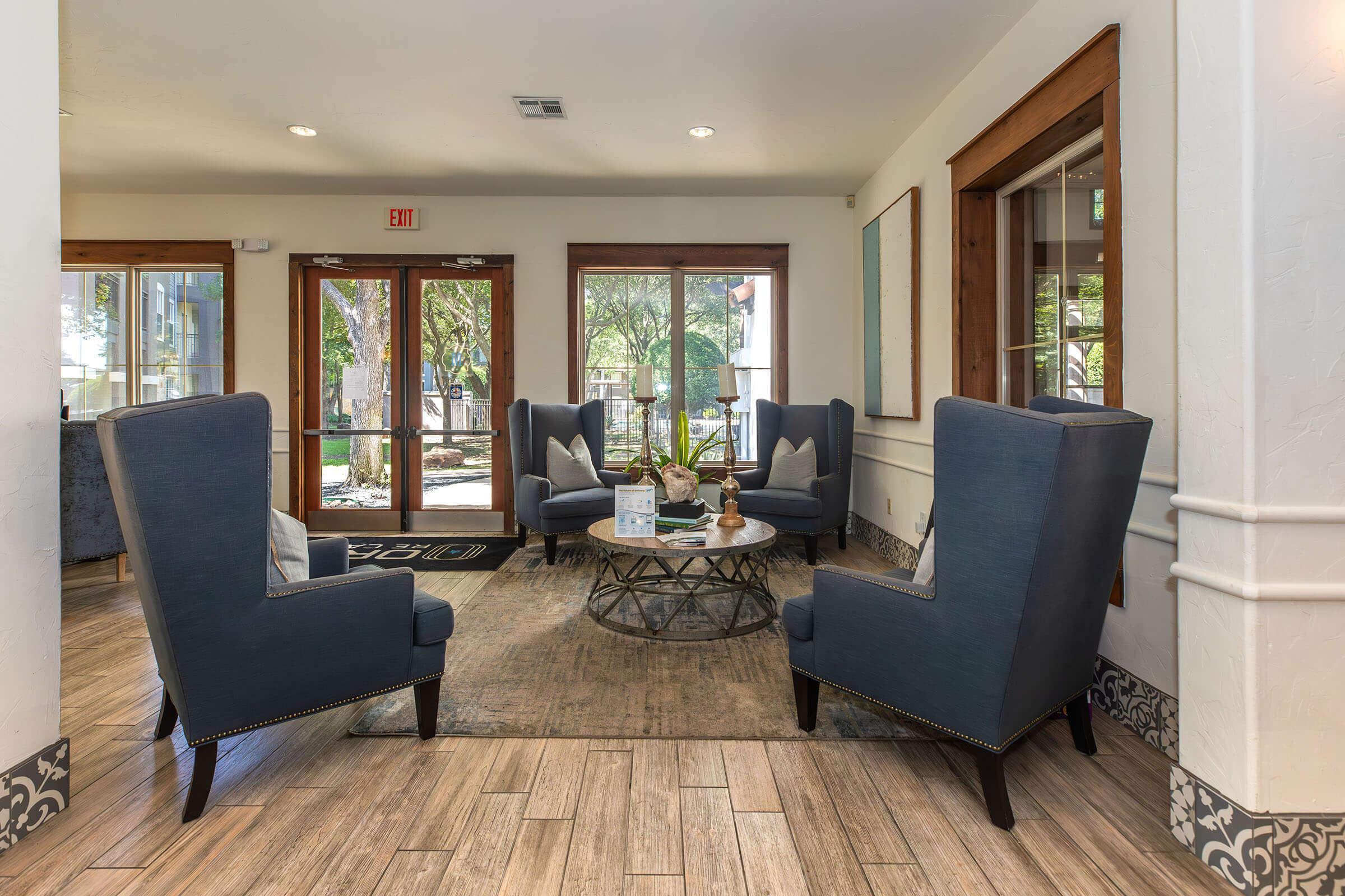 A cozy lounge area featuring two blue wingback chairs and a round coffee table, surrounded by natural light from large windows. The space includes a patterned rug and decorative elements on the table, creating an inviting atmosphere with potted plants visible outside.