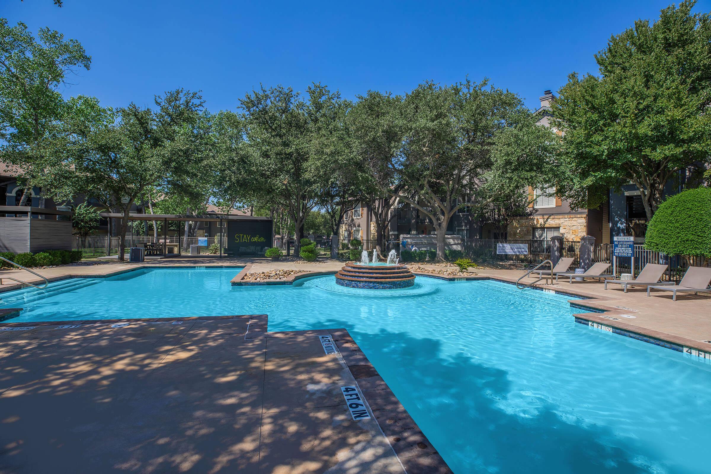 A bright outdoor swimming pool surrounded by lush trees and sun loungers. The pool features a small fountain and is located within a landscaped area, with nearby residential buildings visible in the background. Clear blue skies enhance the inviting atmosphere.