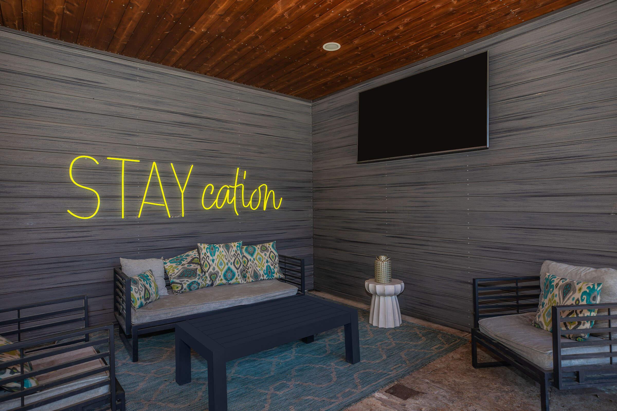 A cozy outdoor seating area with a modern aesthetic. The space features a wooden ceiling and gray wooden walls. There are comfortable sofas with patterned cushions and a coffee table in the center. A television is mounted on the wall, and the phrase "STAYcation" is displayed in bright neon.