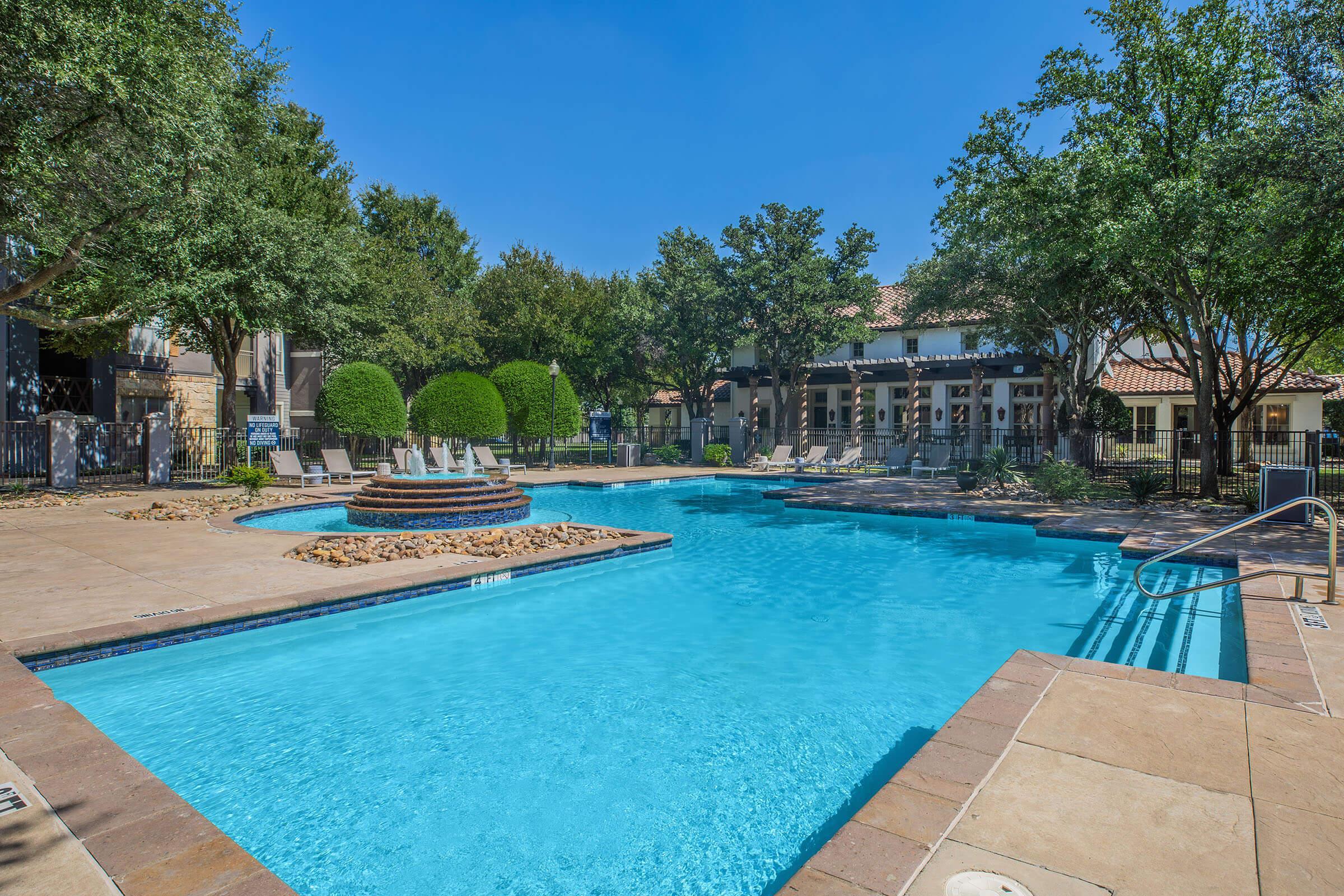 A clear blue swimming pool with a fountain feature surrounded by lounge chairs. Lush greenery and trees are visible in the background, providing a serene outdoor setting. The sky is bright and sunny, enhancing the inviting atmosphere of the pool area.