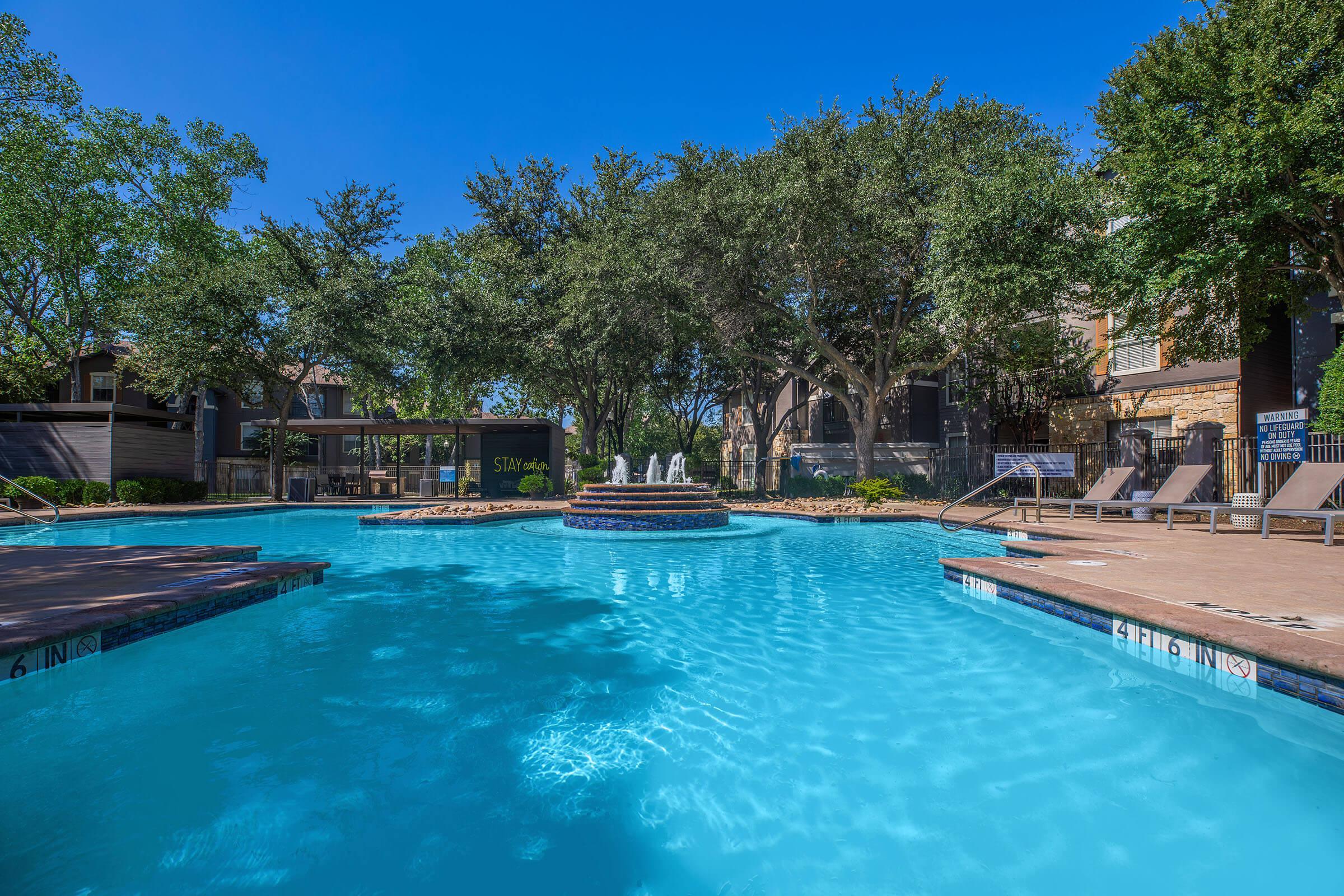 A tranquil swimming pool surrounded by lush green trees, with lounge chairs alongside. In the center, there's a circular fountain creating a relaxing atmosphere. The bright blue sky adds to the serene setting, making it an inviting spot for leisure and relaxation.
