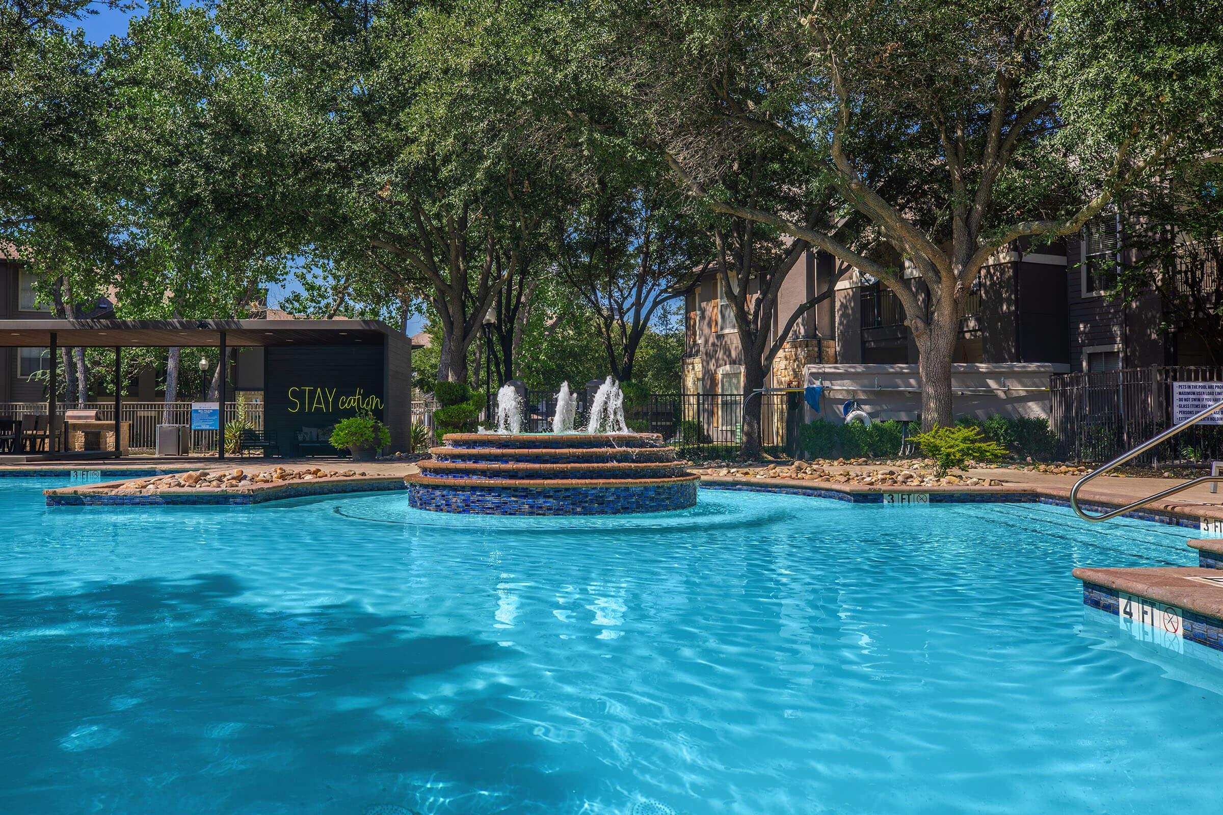 A serene swimming pool surrounded by lush trees and a decorative fountain in the center. In the background, there are shaded areas with outdoor seating and nearby buildings. The water is clear and inviting, reflecting the blue sky above.