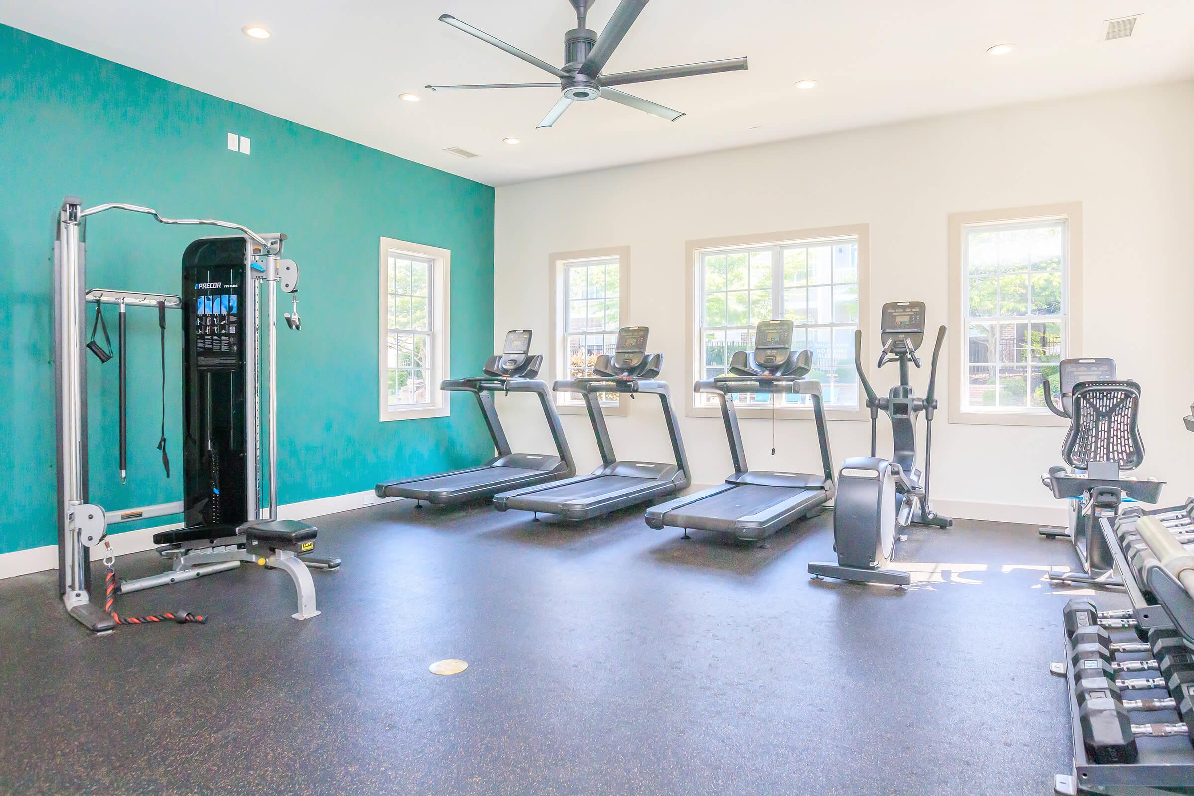 A bright, spacious gym with a turquoise accent wall. Equipped with three treadmills, a weight machine, and a set of free weights on the floor. Large windows provide natural light, enhancing the inviting atmosphere.
