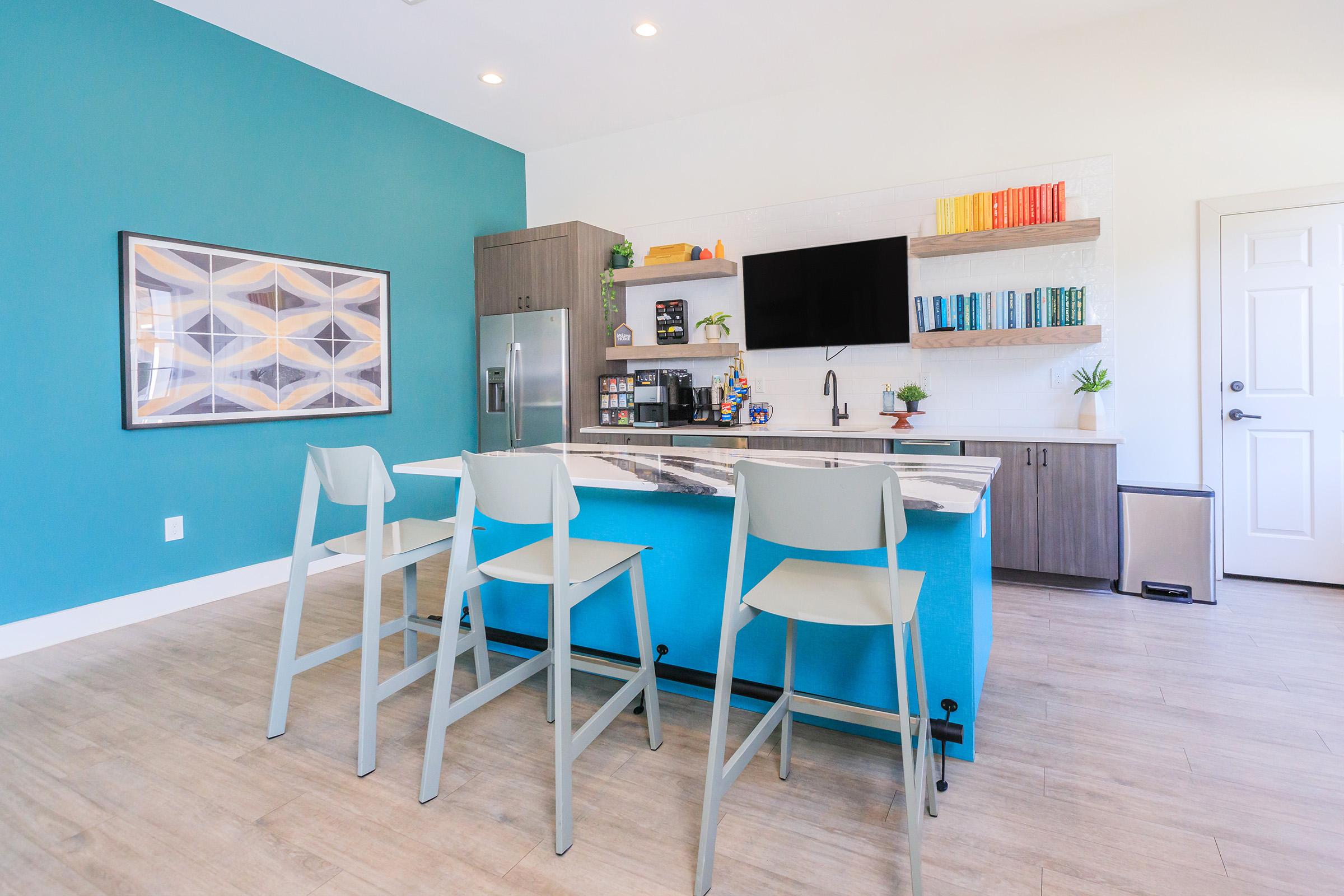 Modern kitchen featuring a vibrant blue accent wall, a central island with bar stools, and wooden cabinetry. The shelves display colorful books and plants, while appliances and a TV provide functionality. Bright lighting enhances the open and airy feel of the space.