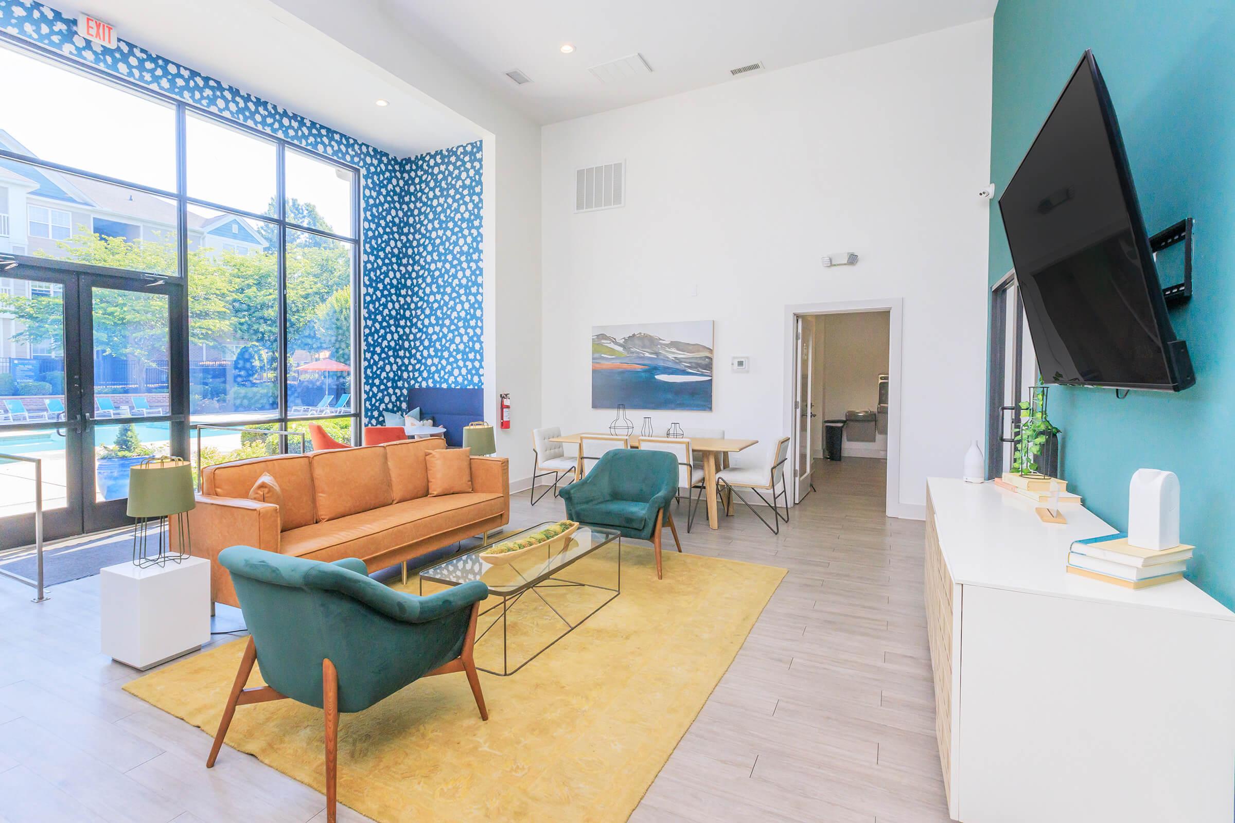 A modern lounge area featuring a bright orange sofa, green armchairs, and a glass coffee table on a yellow rug. Large windows provide natural light and views of the outdoors. Decor includes a contemporary painting and a TV mounted on the wall, with a dining table in the background.