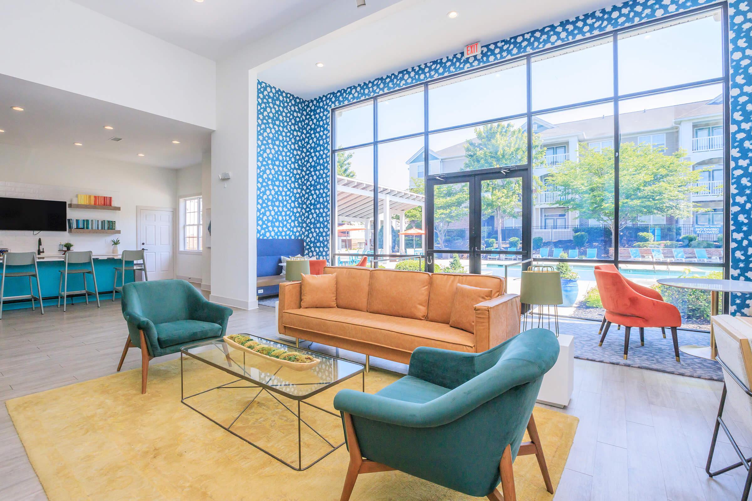 A modern lounge area featuring a large window with outdoor views, a stylish orange couch, teal armchairs, and a glass coffee table. Brightly colored walls and a decorative rug complement the inviting atmosphere, with a glimpse of a pool and greenery outside.