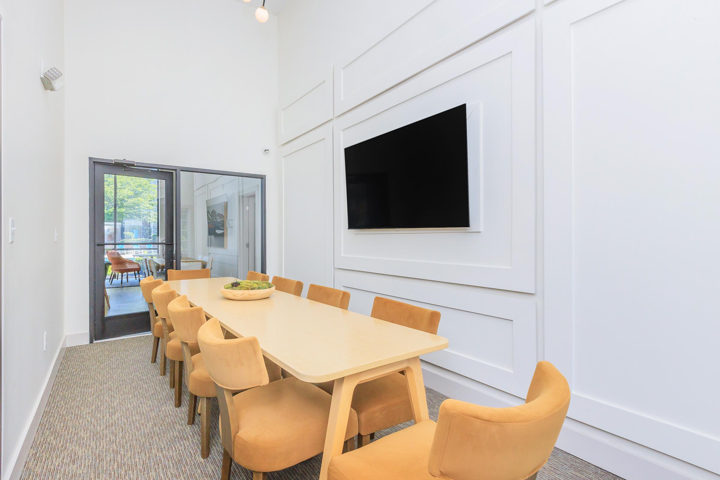 A brightly lit conference room features a long wooden table surrounded by yellow upholstered chairs. One wall includes a large flat-screen TV, while a doorway leads to an outdoor view with additional seating visible. The room is designed with clean lines and neutral tones, creating a modern and inviting atmosphere.