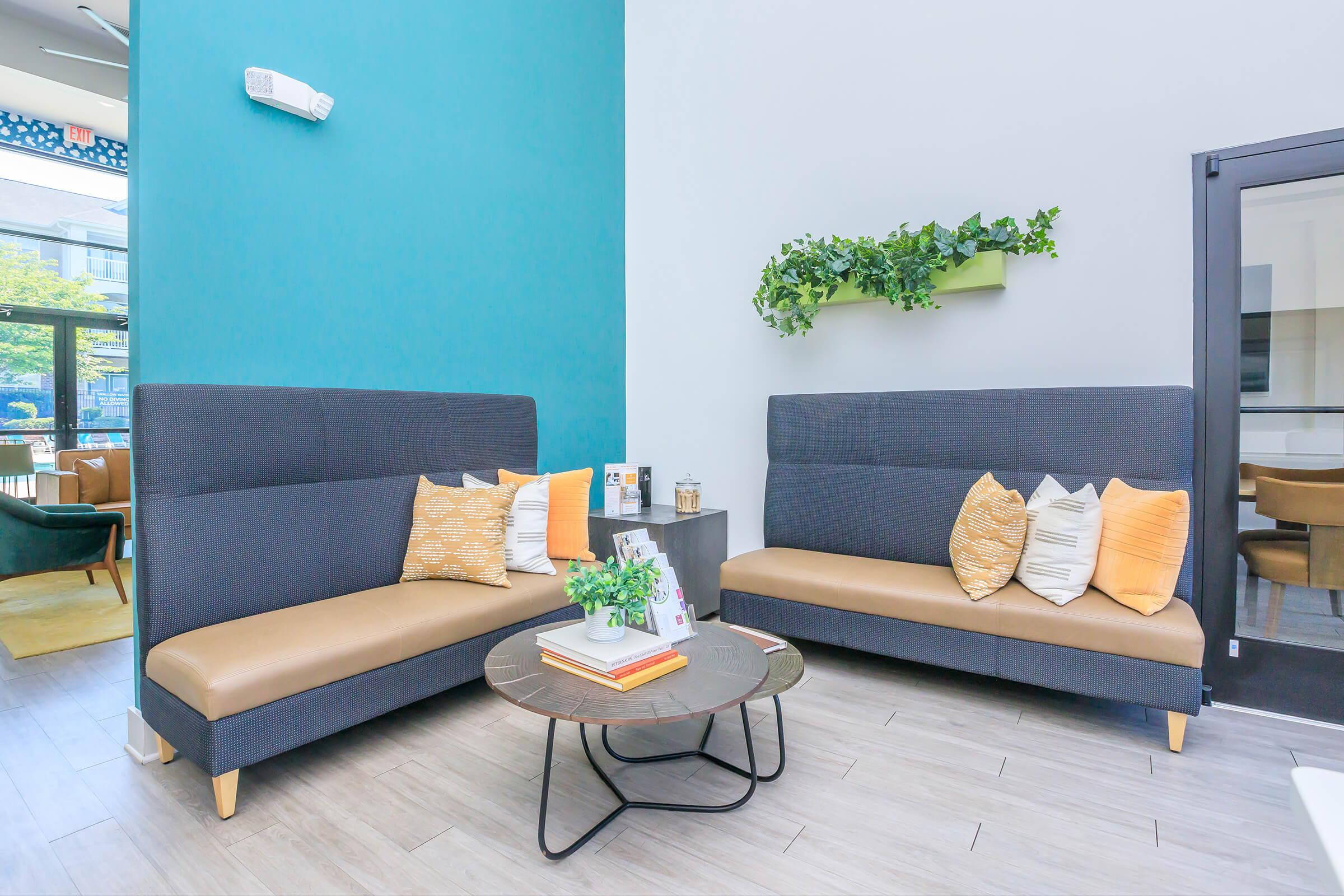 A modern lounge area featuring two dark gray sofas with tan seating and decorative pillows, arranged around a circular coffee table. A small plant sits on the table, and a green wall planter with foliage is mounted on the wall. Natural light comes from large windows in the background.