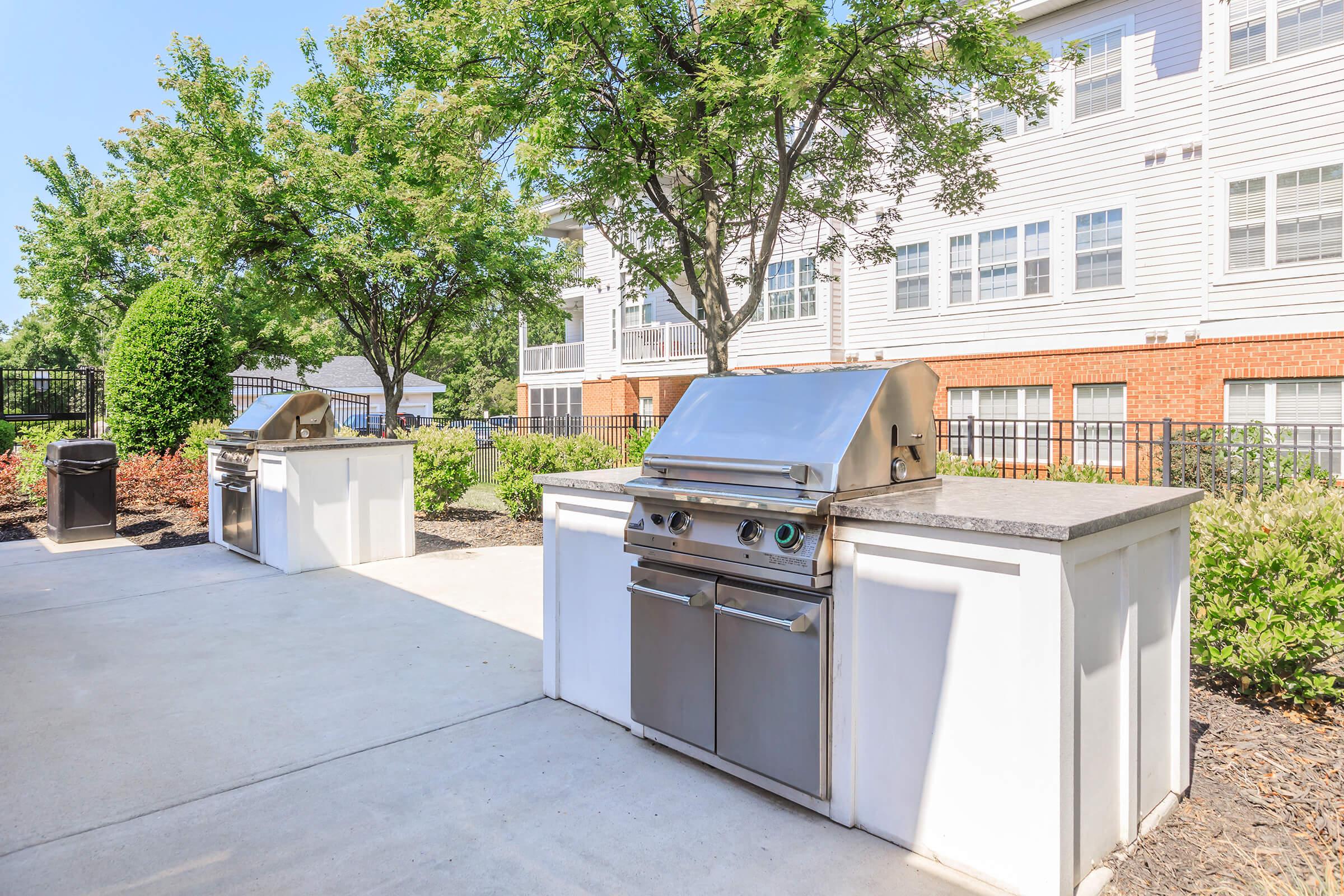 Outdoor barbecue area featuring two stainless steel grills on white stone counters, surrounded by lush greenery and trees. The setting is part of an apartment complex, with a clear sky and well-maintained landscaping. Ideal for outdoor cooking and gatherings.