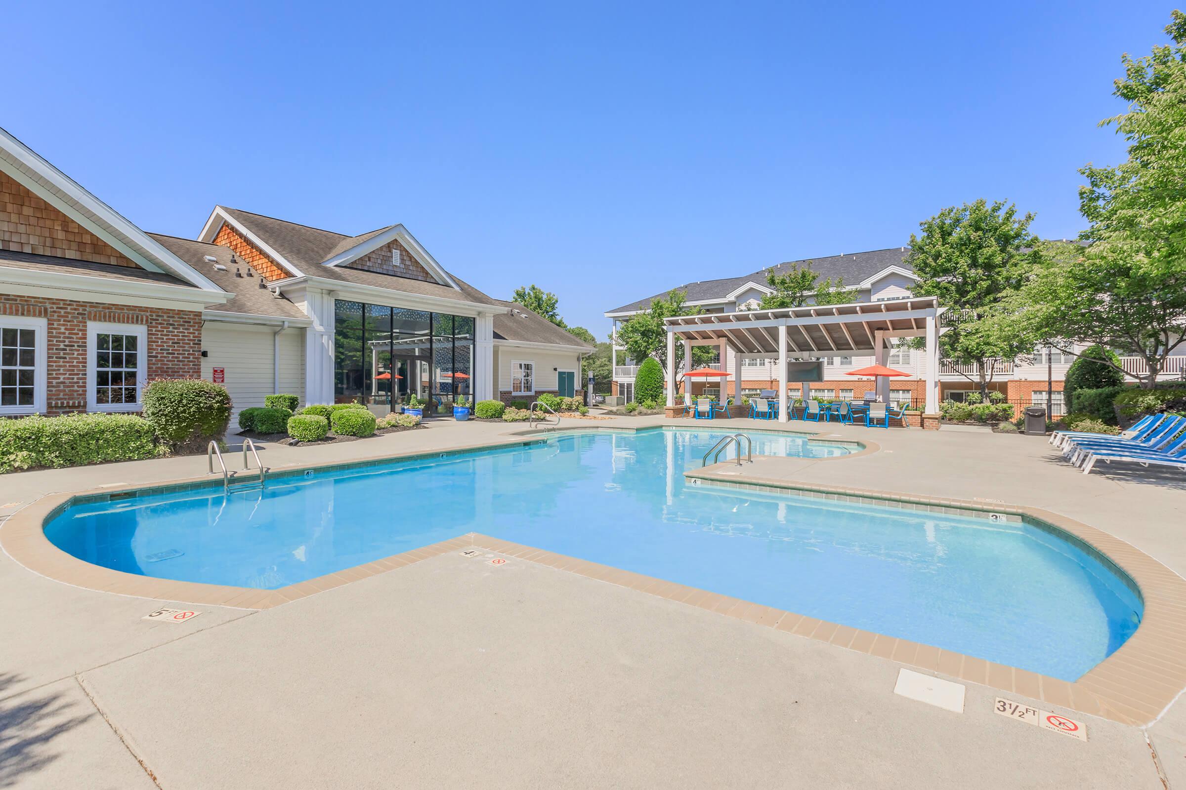 A clean outdoor swimming pool surrounded by patio area, lounge chairs, and greenery. The pool is clear and inviting, with a clubhouse visible in the background under a clear blue sky.