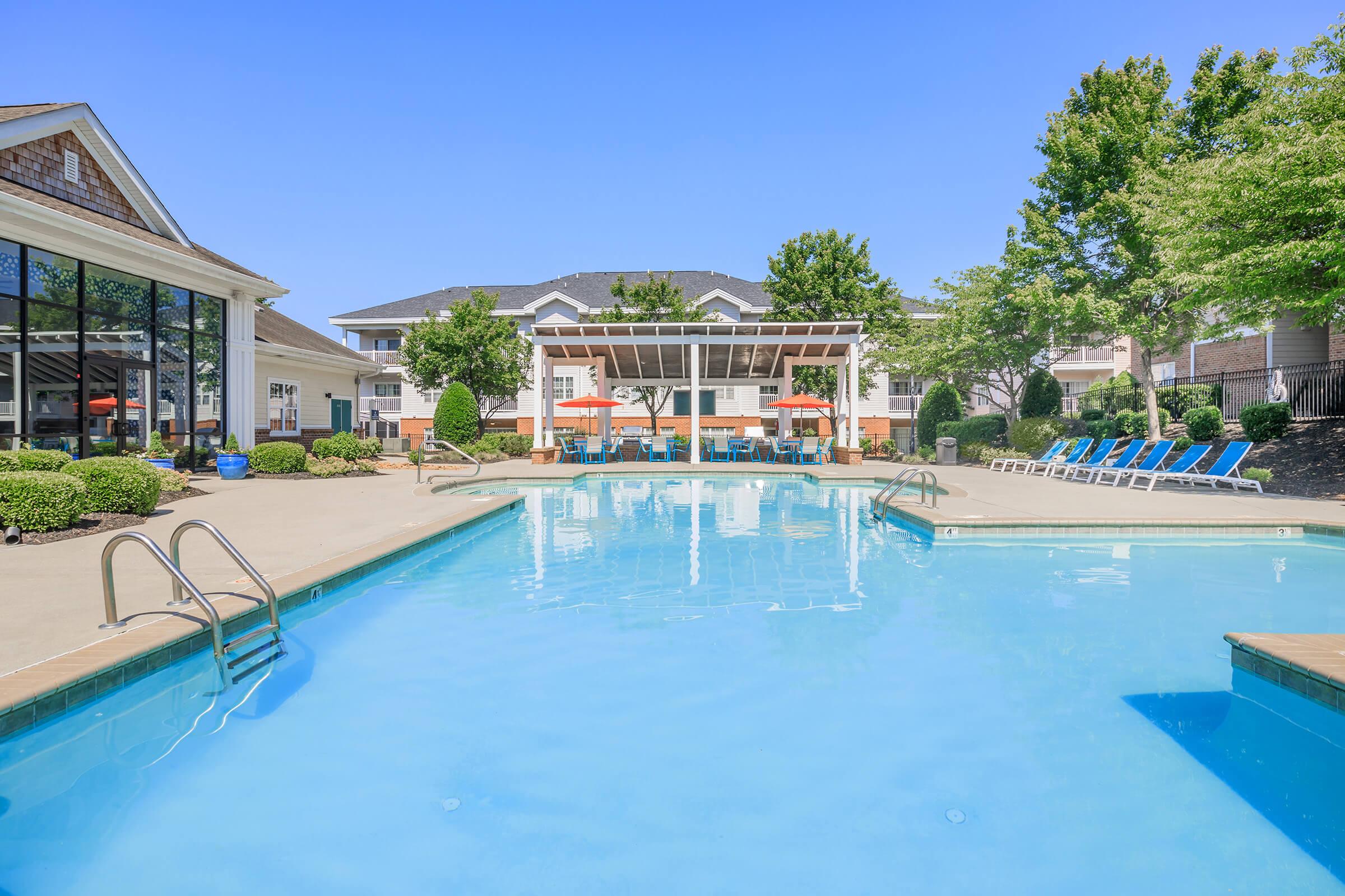 A clear swimming pool surrounded by lounge chairs and neatly trimmed bushes. In the background, a building features shaded seating areas with orange umbrellas. The sky is bright blue, creating a cheerful and inviting atmosphere for relaxation and leisure.