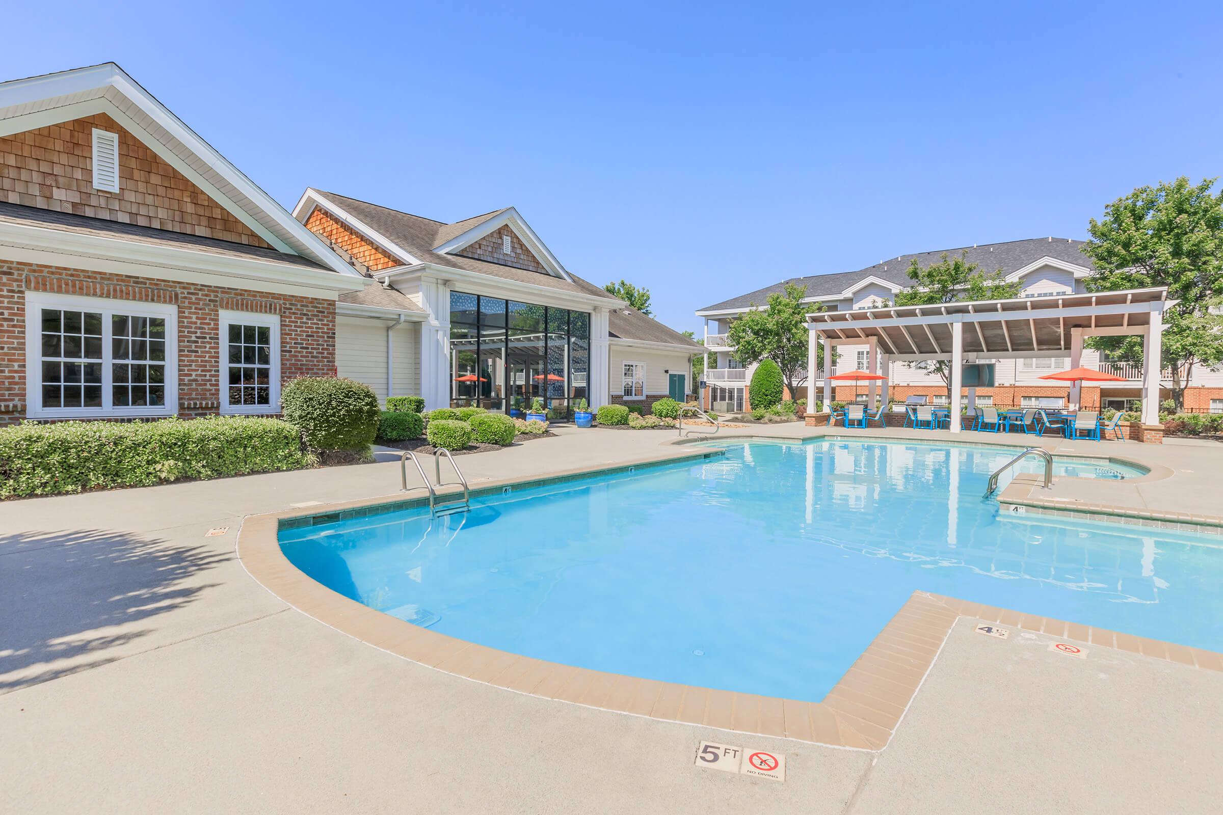 A bright and inviting outdoor pool area with clear blue water, surrounded by neatly landscaped greenery. Visible are lounge chairs, a shaded seating area with tables, and a building with large windows in the background, all under a clear blue sky.