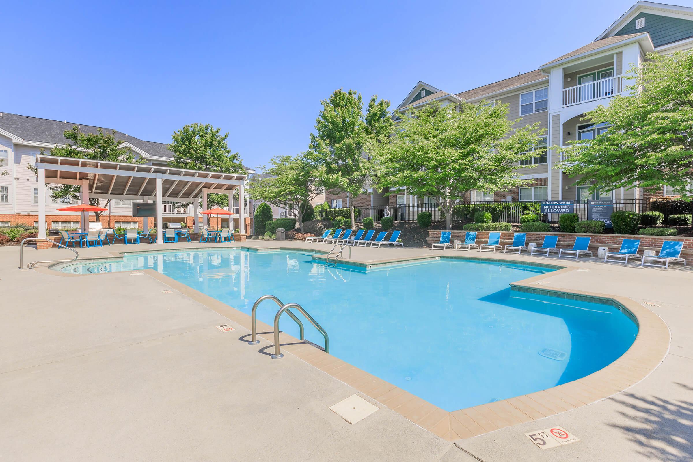 A clear, inviting swimming pool surrounded by lounge chairs and greenery. The pool area features a shaded cabana and is situated near modern apartment buildings under a blue sky. The setting suggests a relaxing and recreational atmosphere.