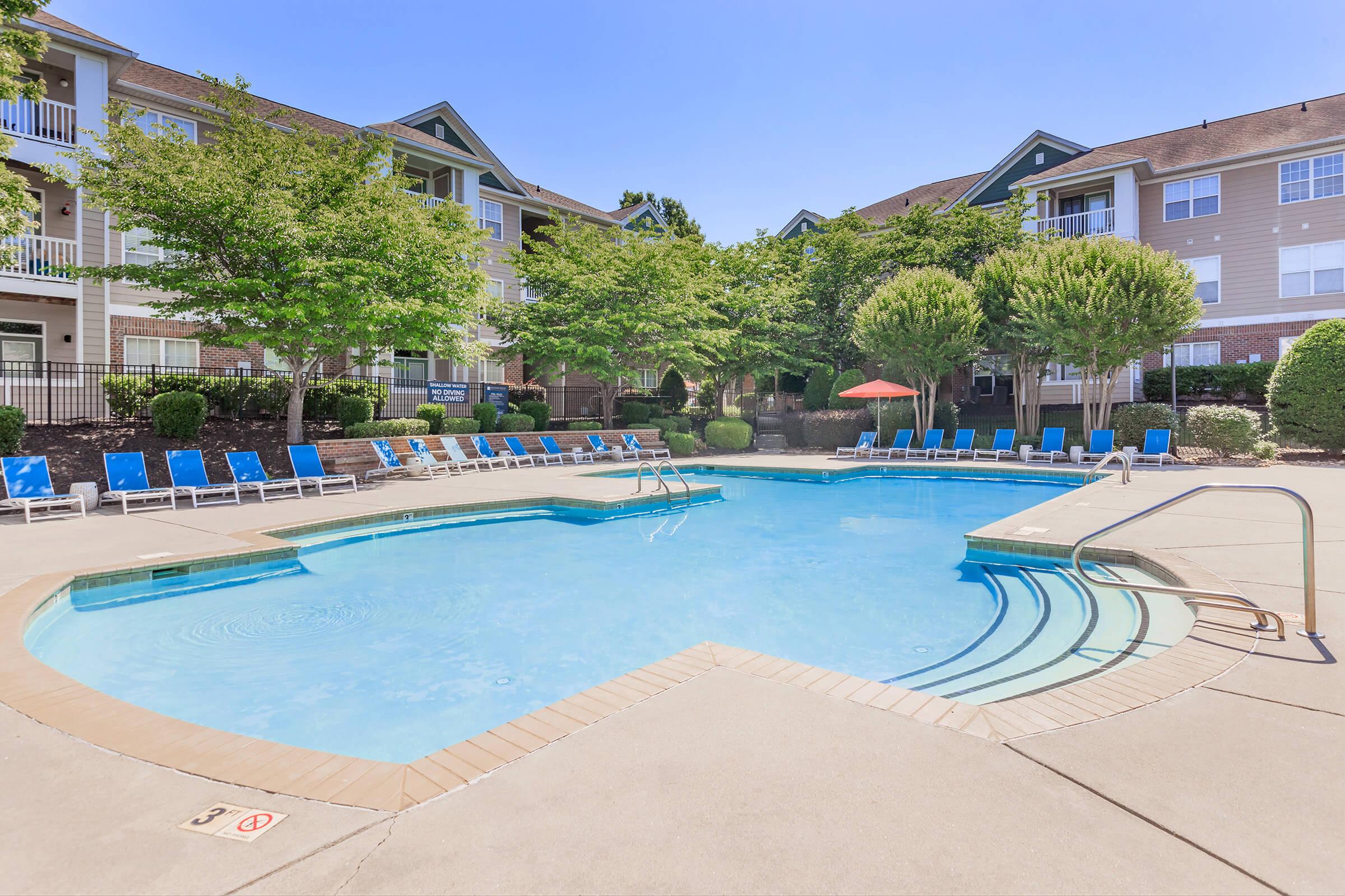 A clear blue swimming pool surrounded by lounge chairs and shaded areas, set in a landscaped area with trees and apartment buildings in the background. The scene is bathed in sunlight, creating a relaxing atmosphere ideal for leisure and recreation.