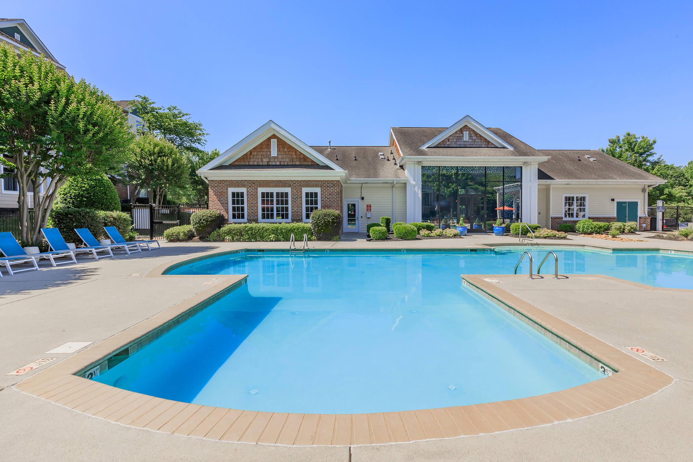 A serene swimming pool surrounded by lounge chairs, with a modern clubhouse in the background. The pool features two sections, and the area is well-maintained with greenery and clear blue skies overhead, creating a relaxing atmosphere.