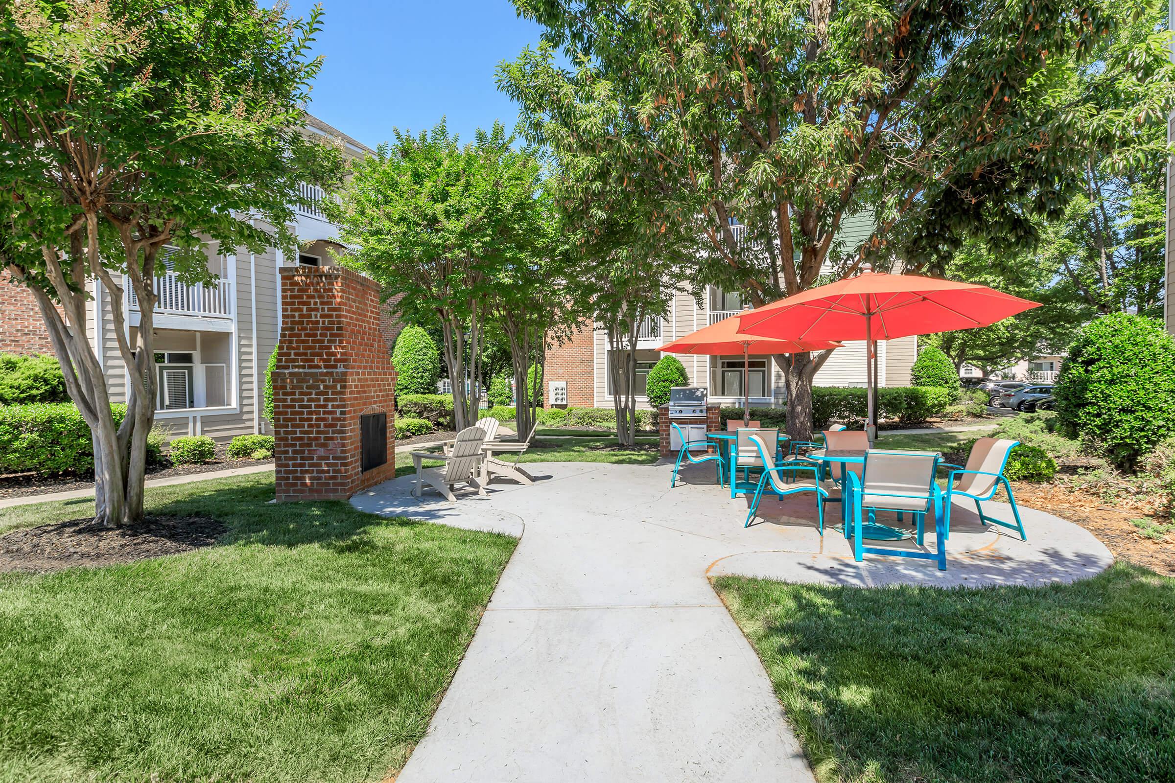 A landscaped outdoor area featuring a paved path surrounded by greenery, with several chairs and a vibrant red umbrella over a table. There is also a brick outdoor fireplace and trees providing shade, creating a relaxing atmosphere in a residential complex.