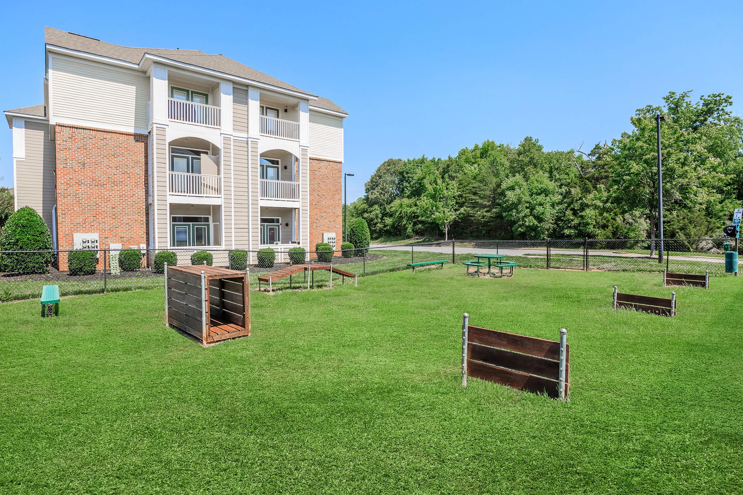 A green outdoor space with a fenced area, featuring a grassy lawn, picnic tables, and wooden structures. In the background, there is a beige and brick apartment building under a clear blue sky. The setting appears to be designed for leisure or recreational activities.